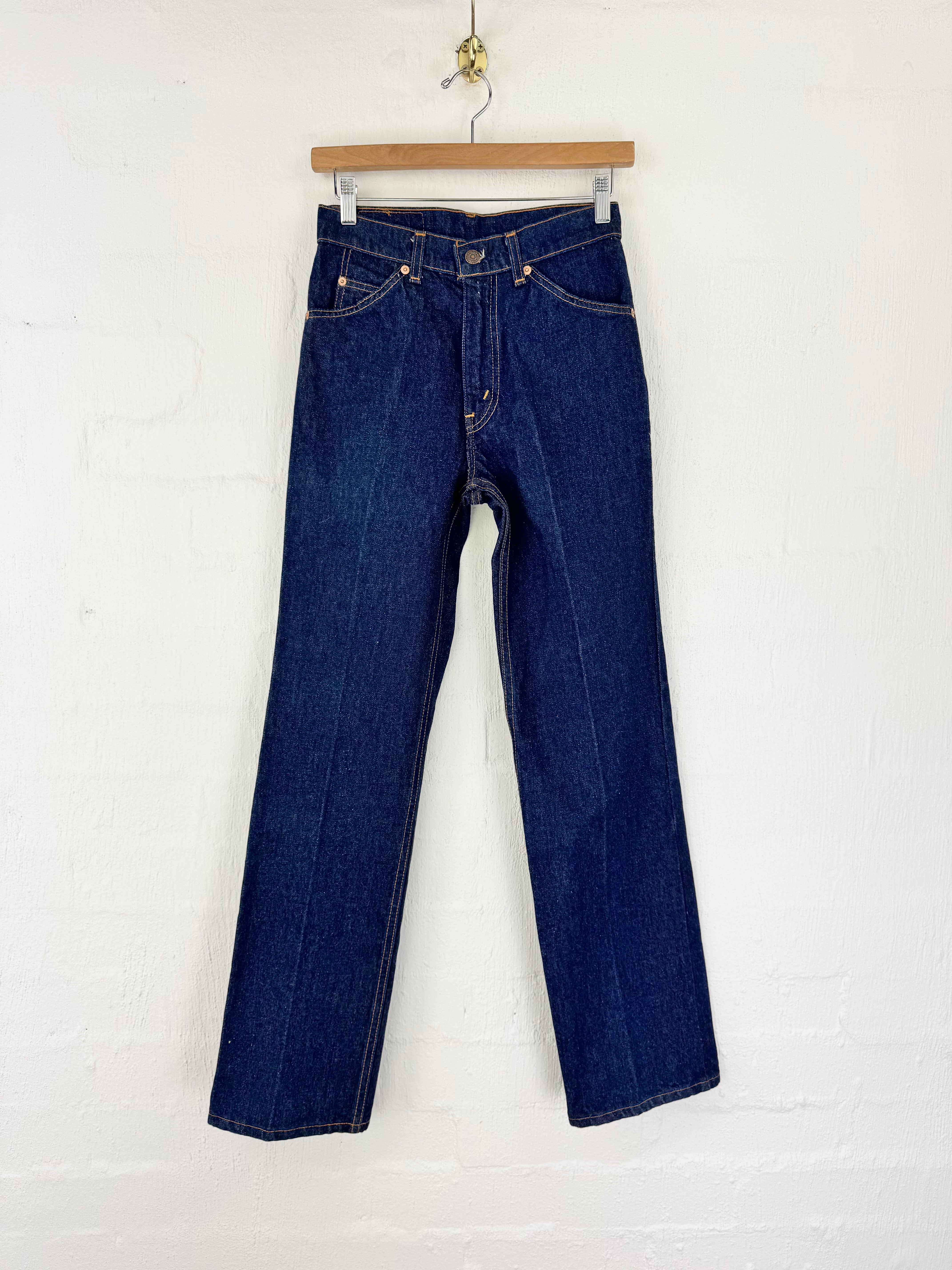 A pair of dark blue denim jeans hanging from a wooden hanger against a white painted brick wall.