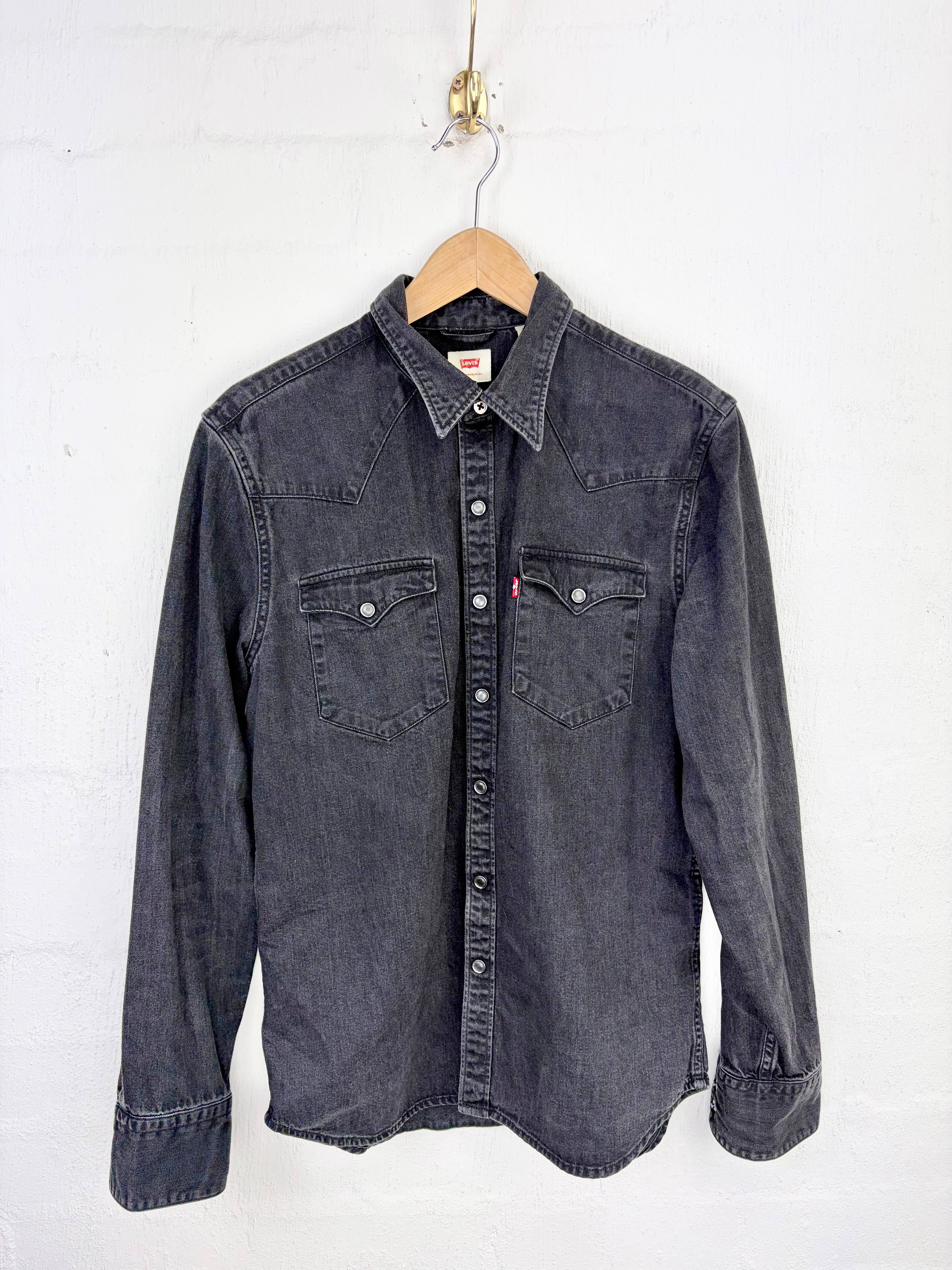 A dark gray denim western shirt jacket hanging on a wooden hanger against a white painted brick wall.