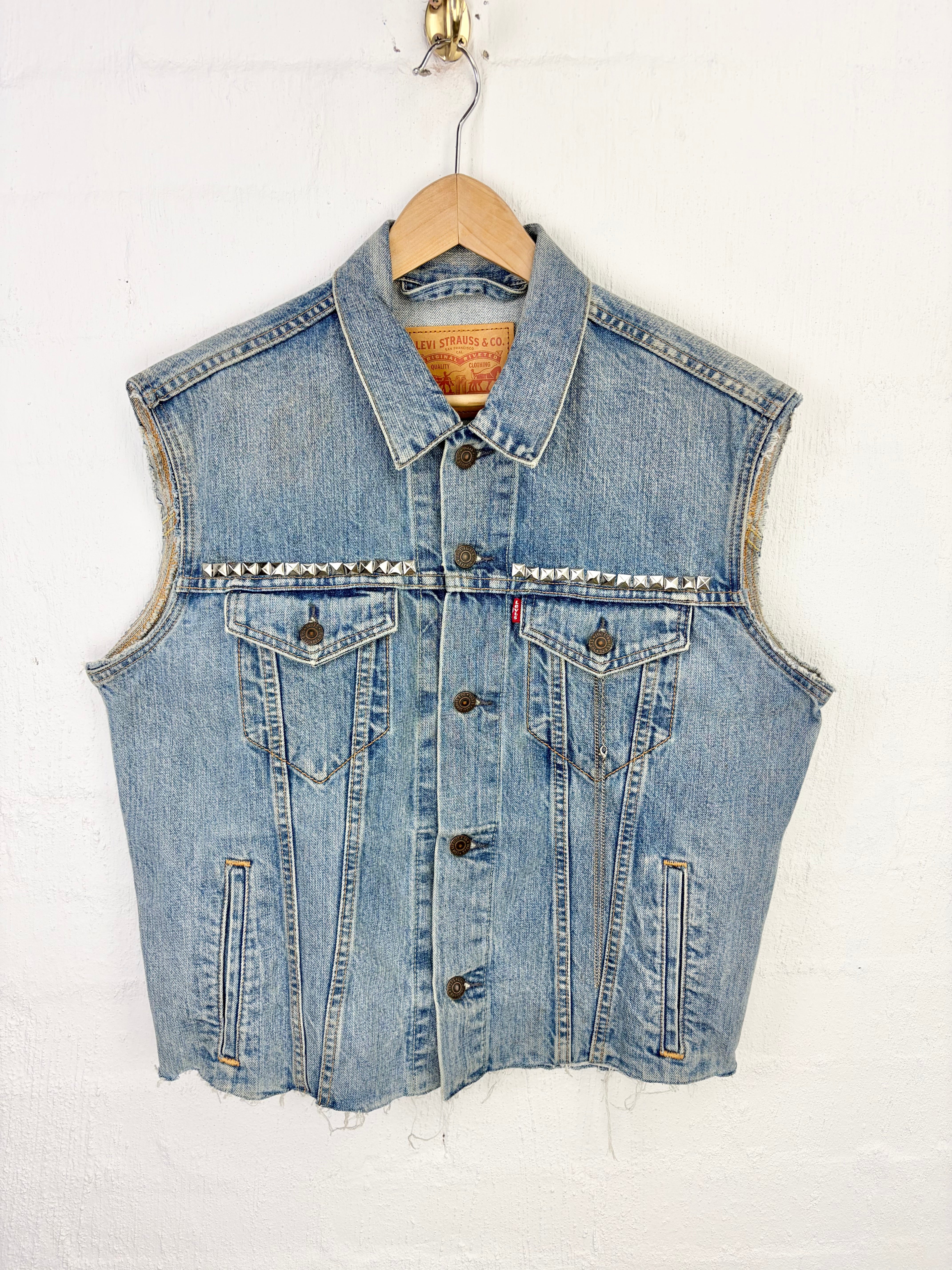 A light blue denim vest with decorative silver studs across the chest, hanging on a wooden hanger against a white textured wall.