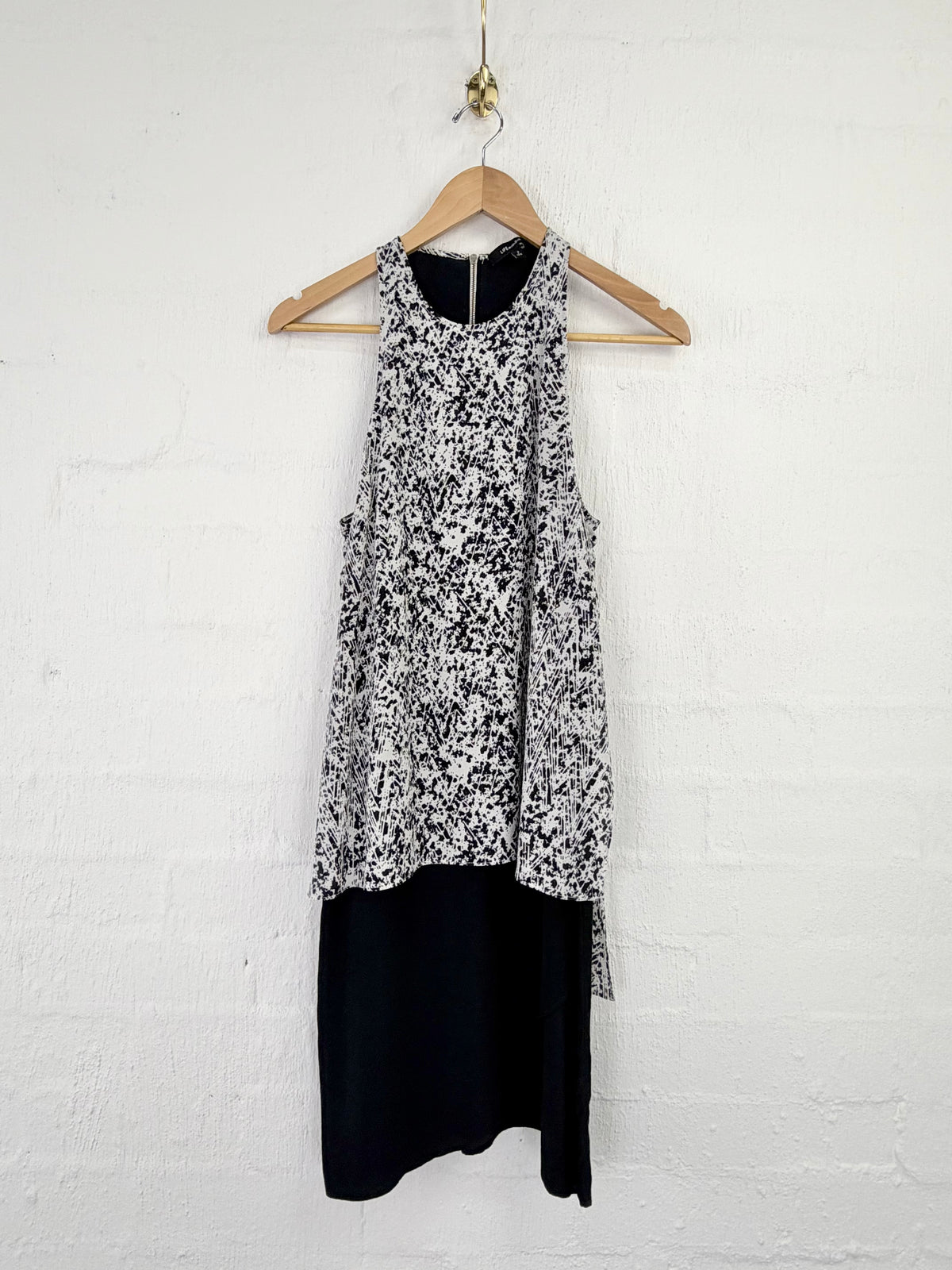 A sleeveless black and white patterned top layered over a solid black skirt, hanging on a wooden hanger against a white painted brick wall.
