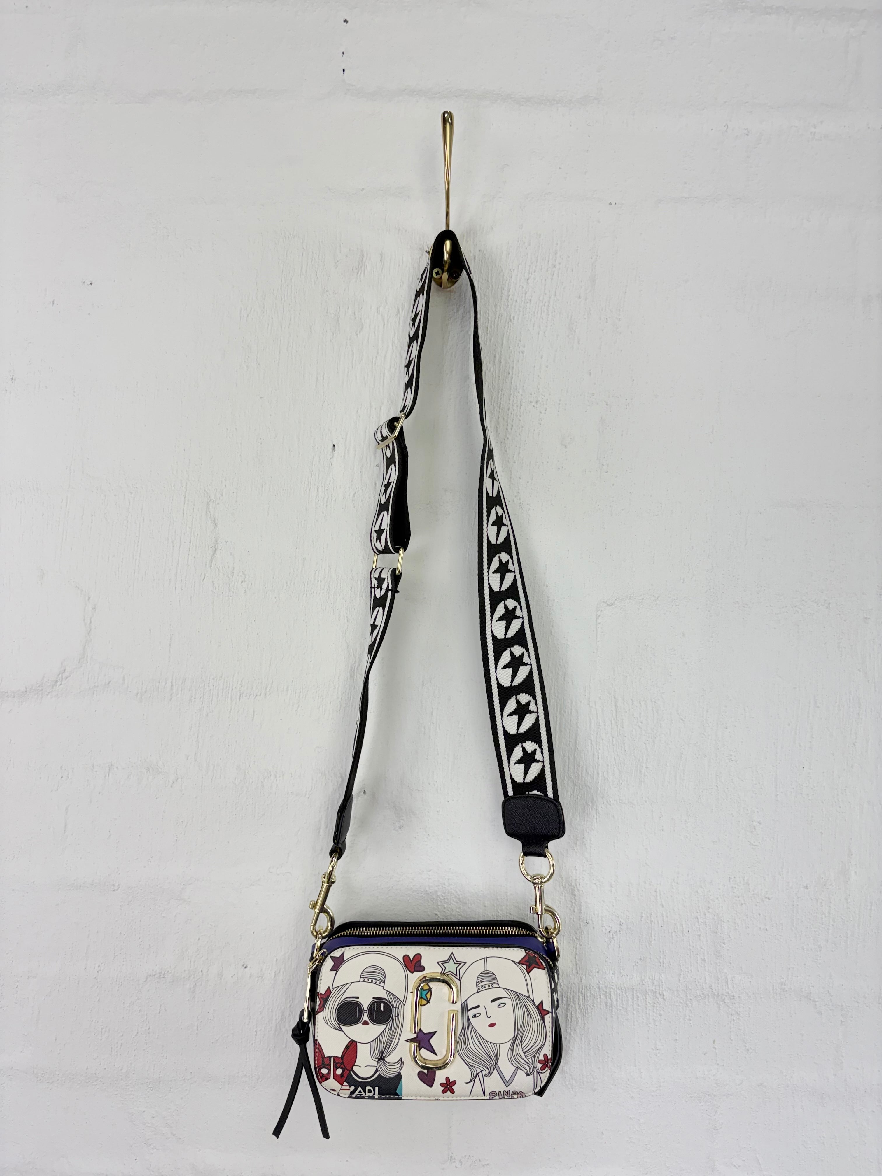 A small crossbody bag with a black and white patterned strap and a printed front pocket featuring cartoon characters, hanging from a gold hook on a white brick wall.