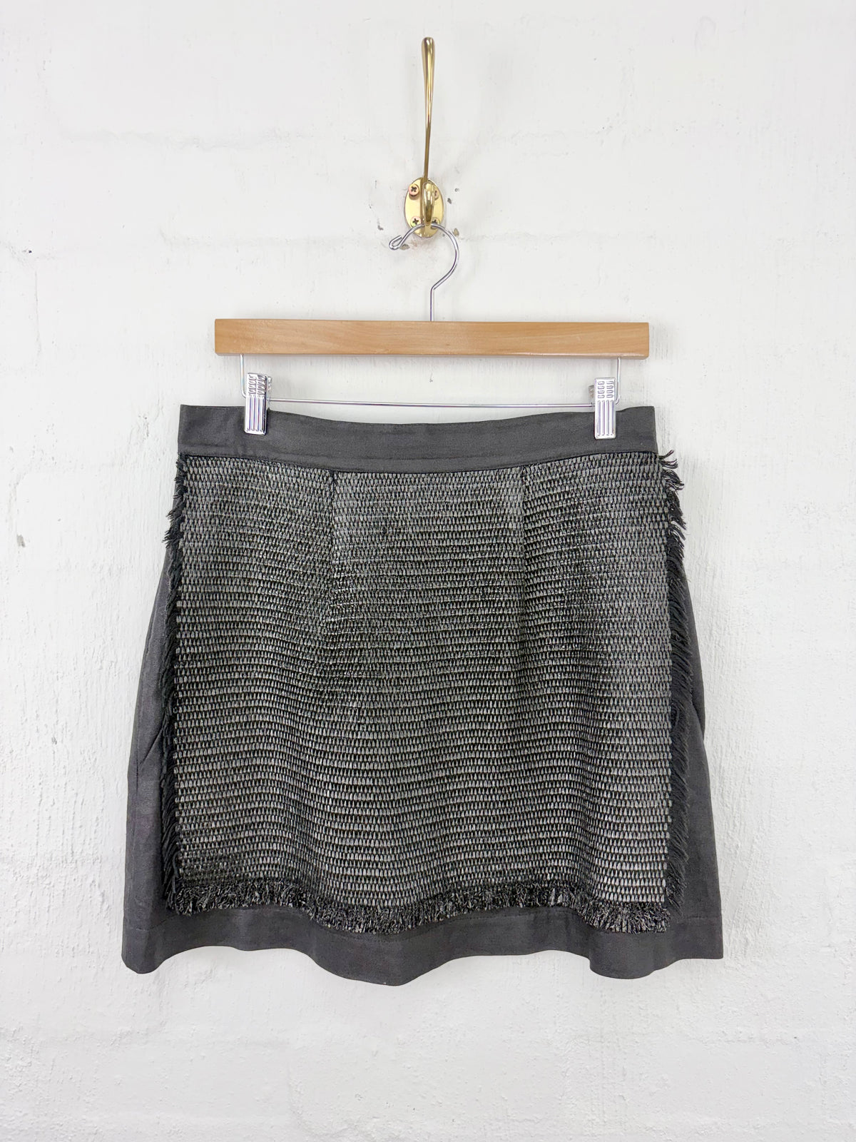 A dark gray textured tweed mini skirt with fringed edges, hanging on a wooden hanger against a white brick wall.