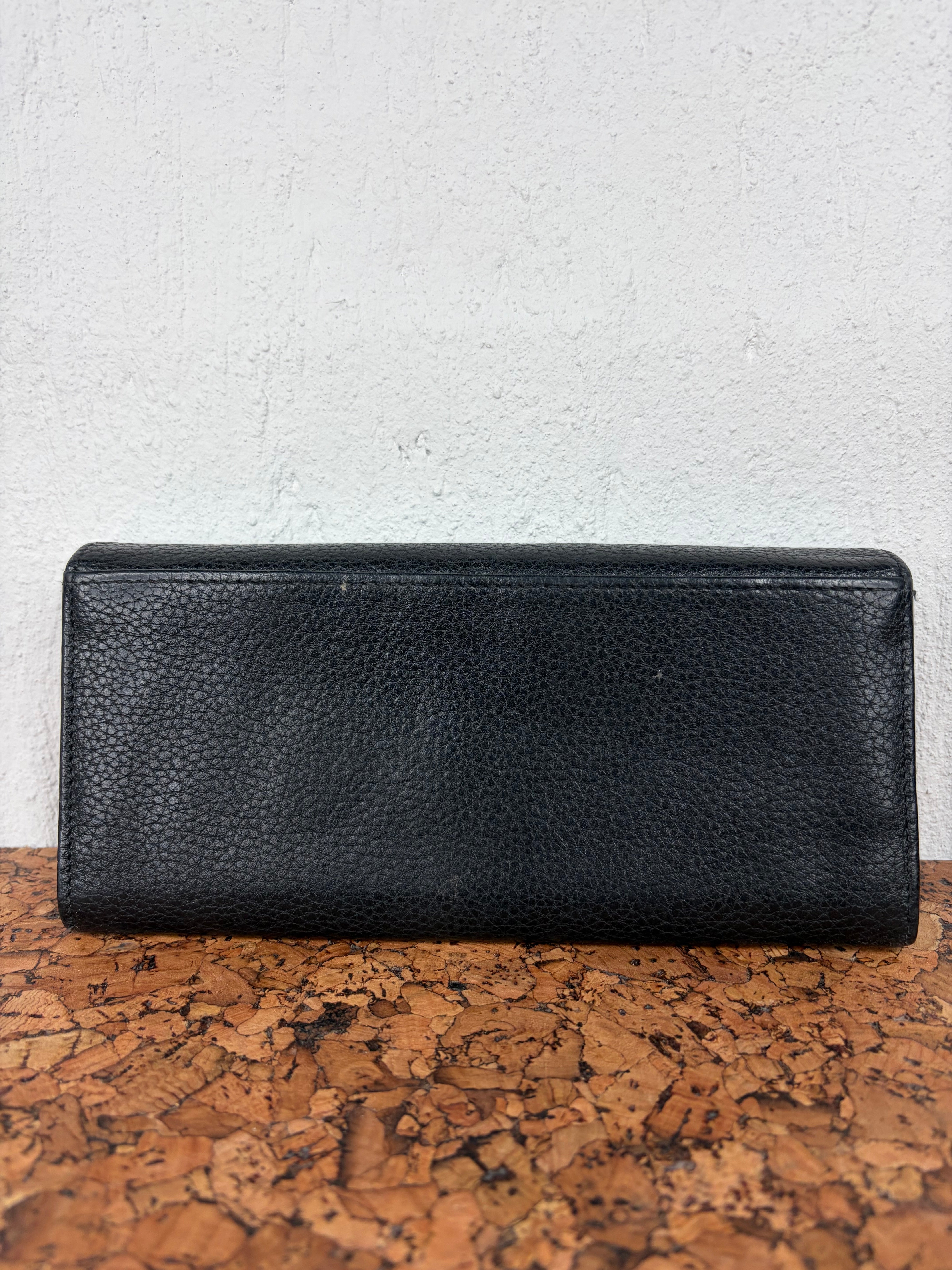 A black pebbled leather long wallet or clutch resting on a textured cork surface against a white wall.