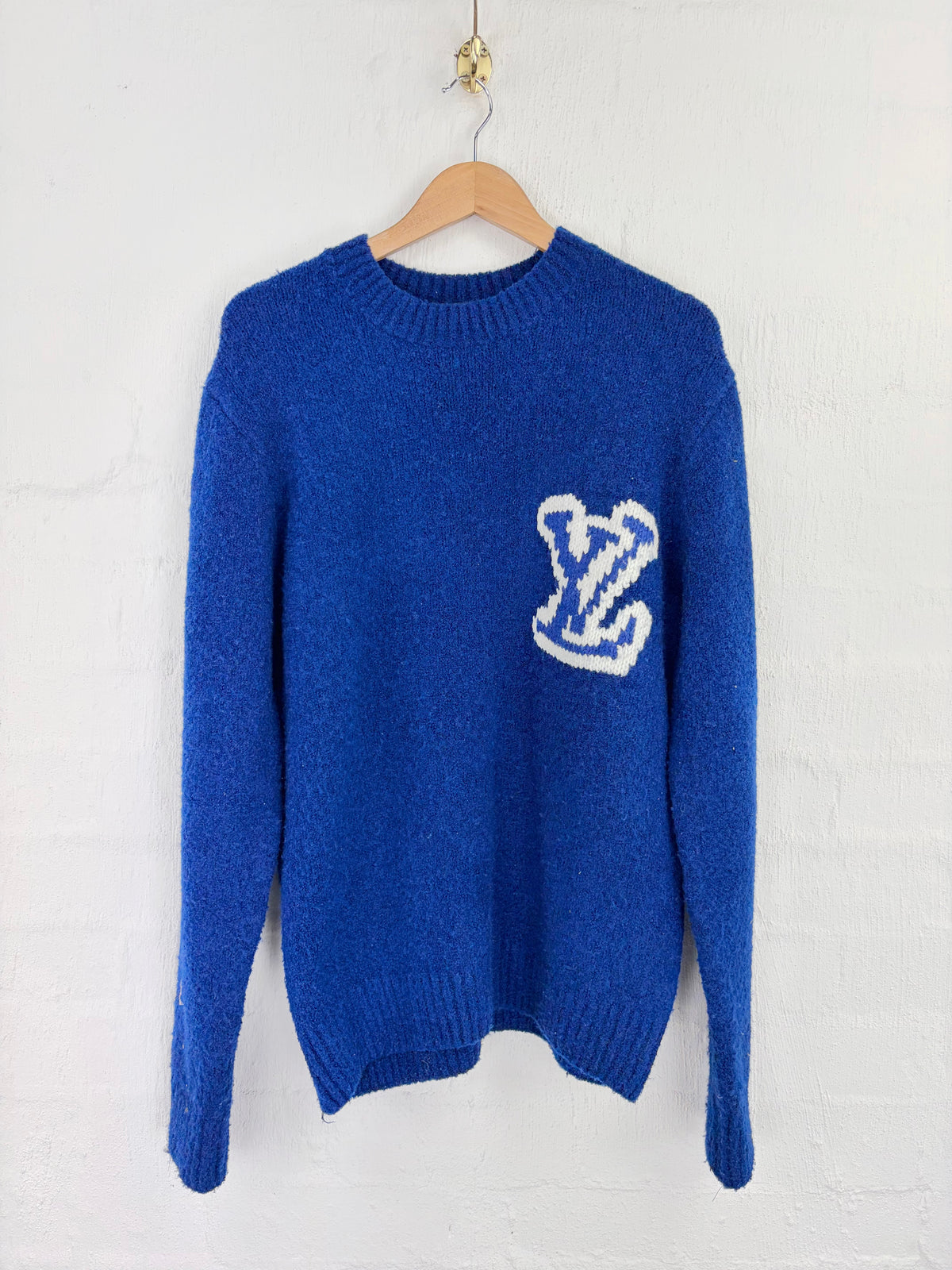 A royal blue knit sweater hanging on a wooden hanger against a white painted brick wall. A white knitted 'LV' monogram is featured on the left chest.