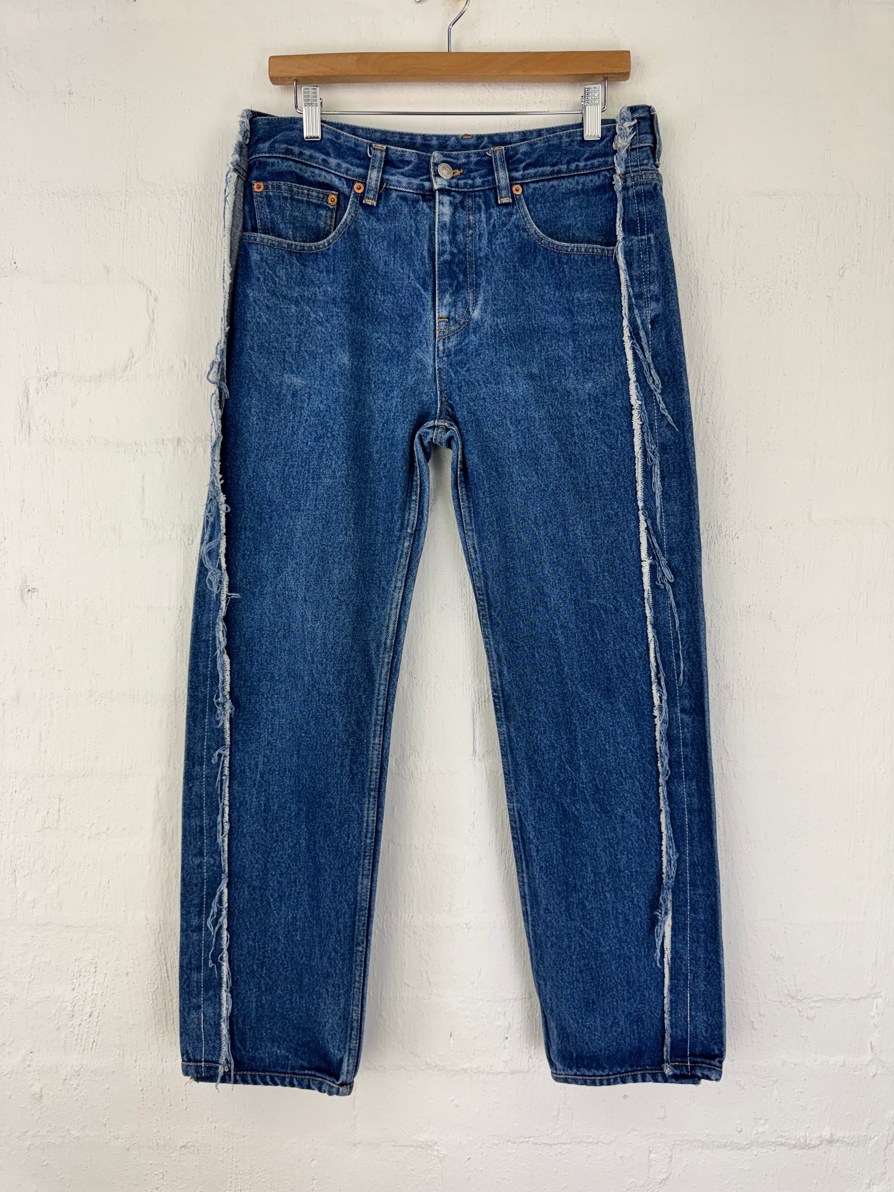 A pair of blue denim jeans with raw, frayed hems and contrast white stitching, hanging flat on a wooden hanger against a white painted brick wall.