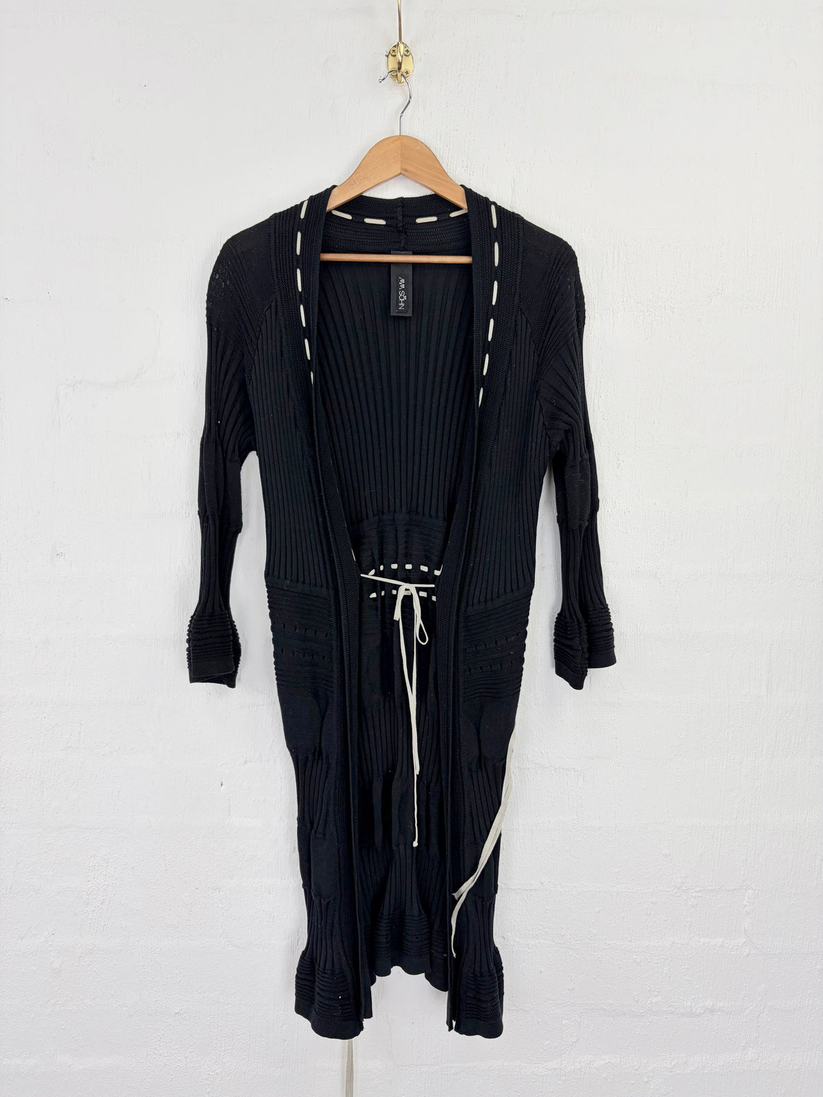A black ribbed knit long cardigan dress with white contrast stitching and a white drawstring belt, hanging on a wooden hanger against a white brick wall.