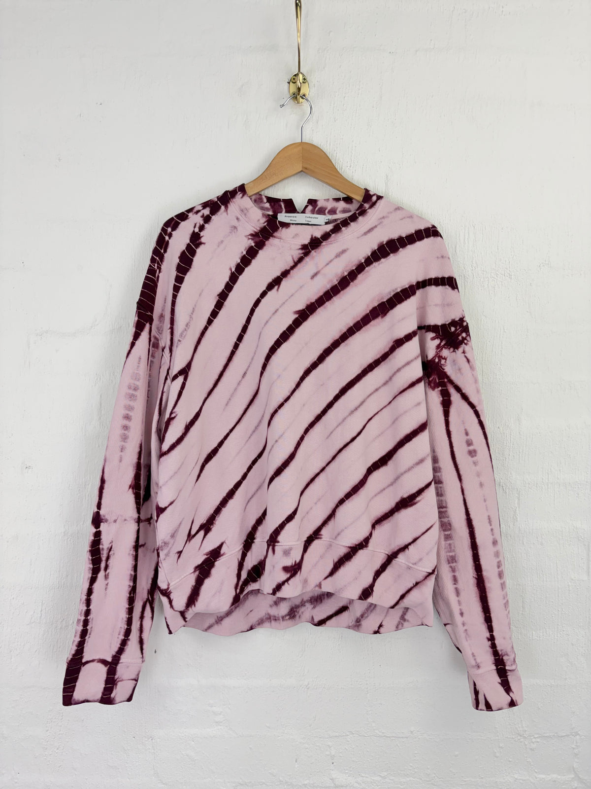 A pink long-sleeve top with a dark purple tie-dye pattern, hanging on a wooden hanger against a white brick wall.