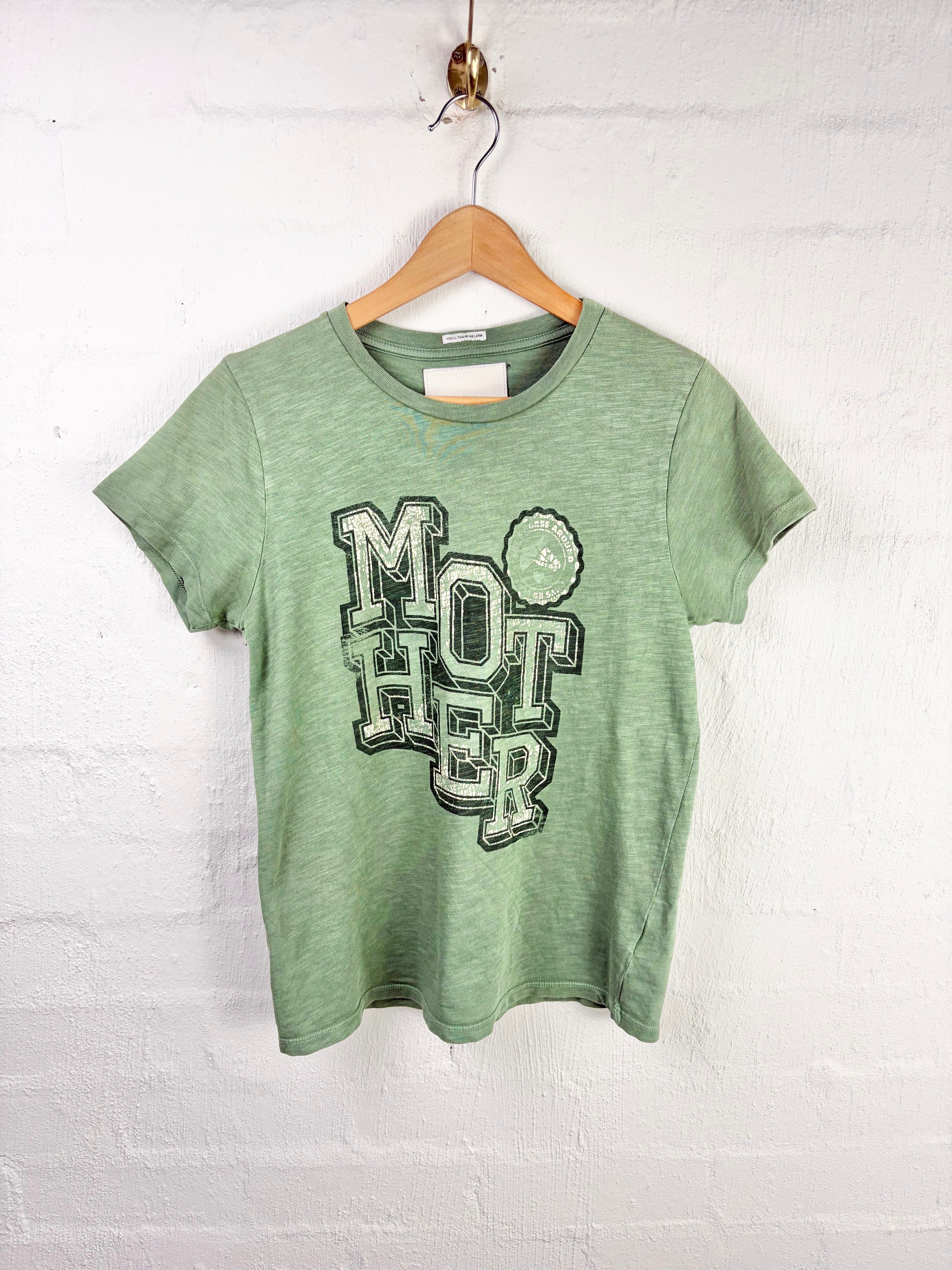 A heather green short-sleeve t-shirt hanging on a wooden hanger against a white painted brick wall. The shirt features a distressed graphic on the front reading 'MOTHER' in dark green block letters with a small green flower detail.