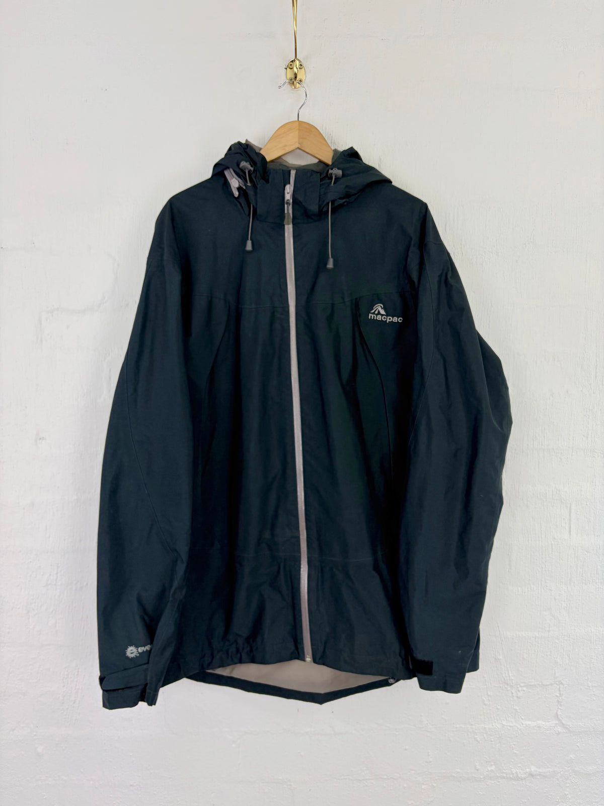 A dark navy hooded rain jacket hanging on a wooden hanger against a white painted brick wall. The jacket features a silver-gray front zipper, a small white logo on the left chest, and a gray inner lining visible at the hem.