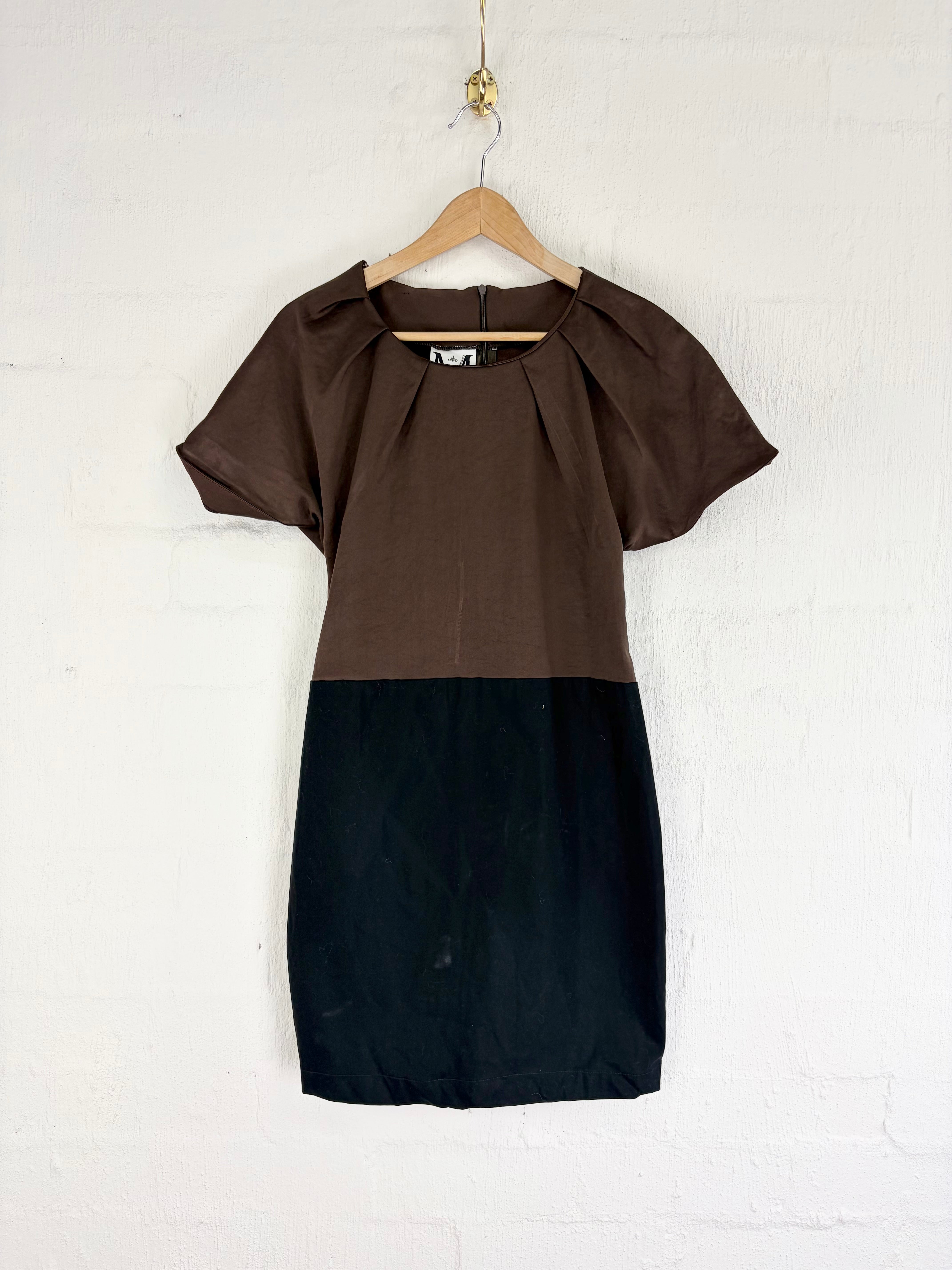 A color-block mini dress with a brown upper bodice and sleeves, and a black lower skirt, hanging on a wooden hanger against a white painted brick wall.