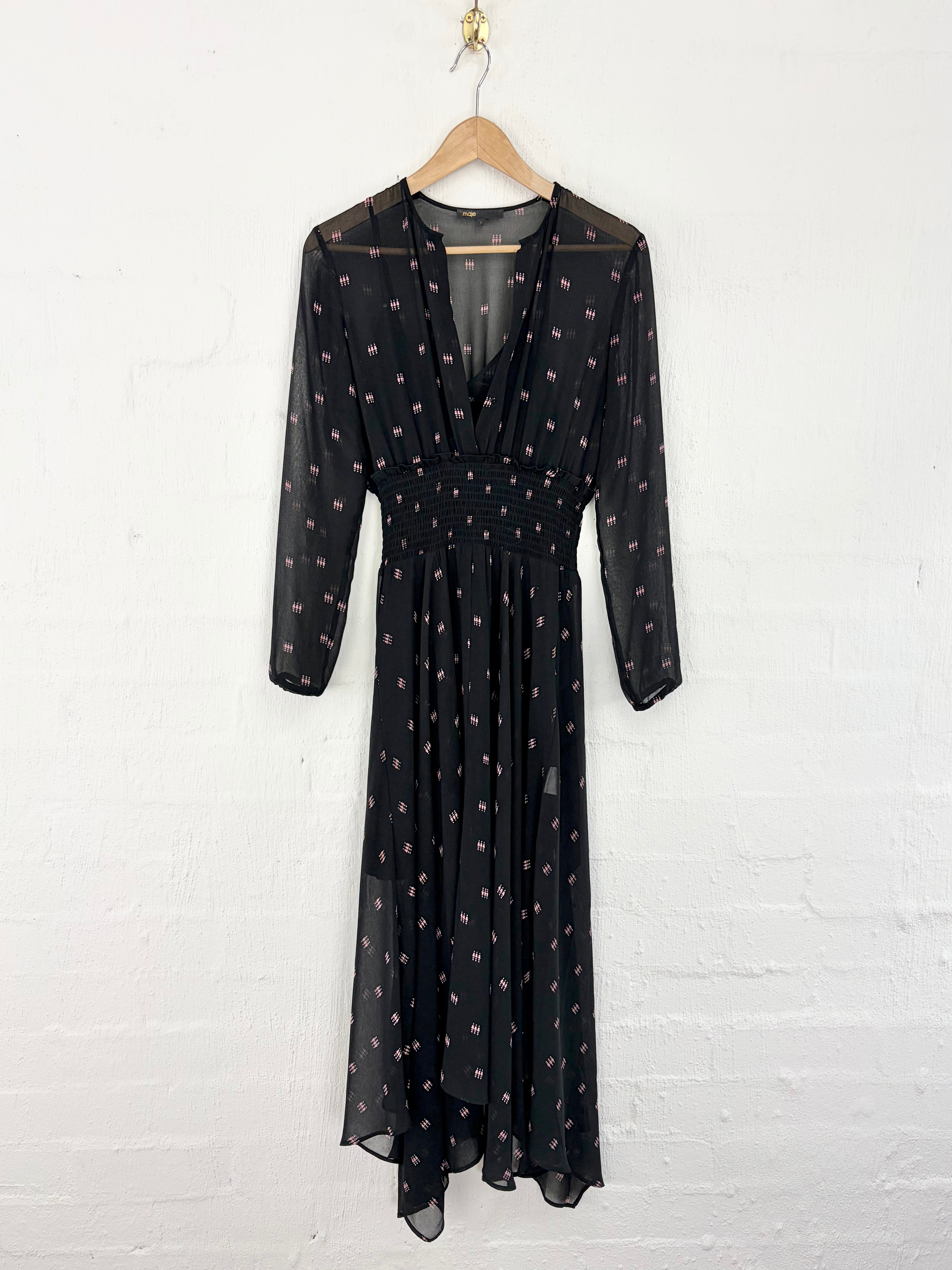 A black sheer midi dress with a small pink and gold polka dot pattern, hanging on a wooden hanger against a white painted brick wall.