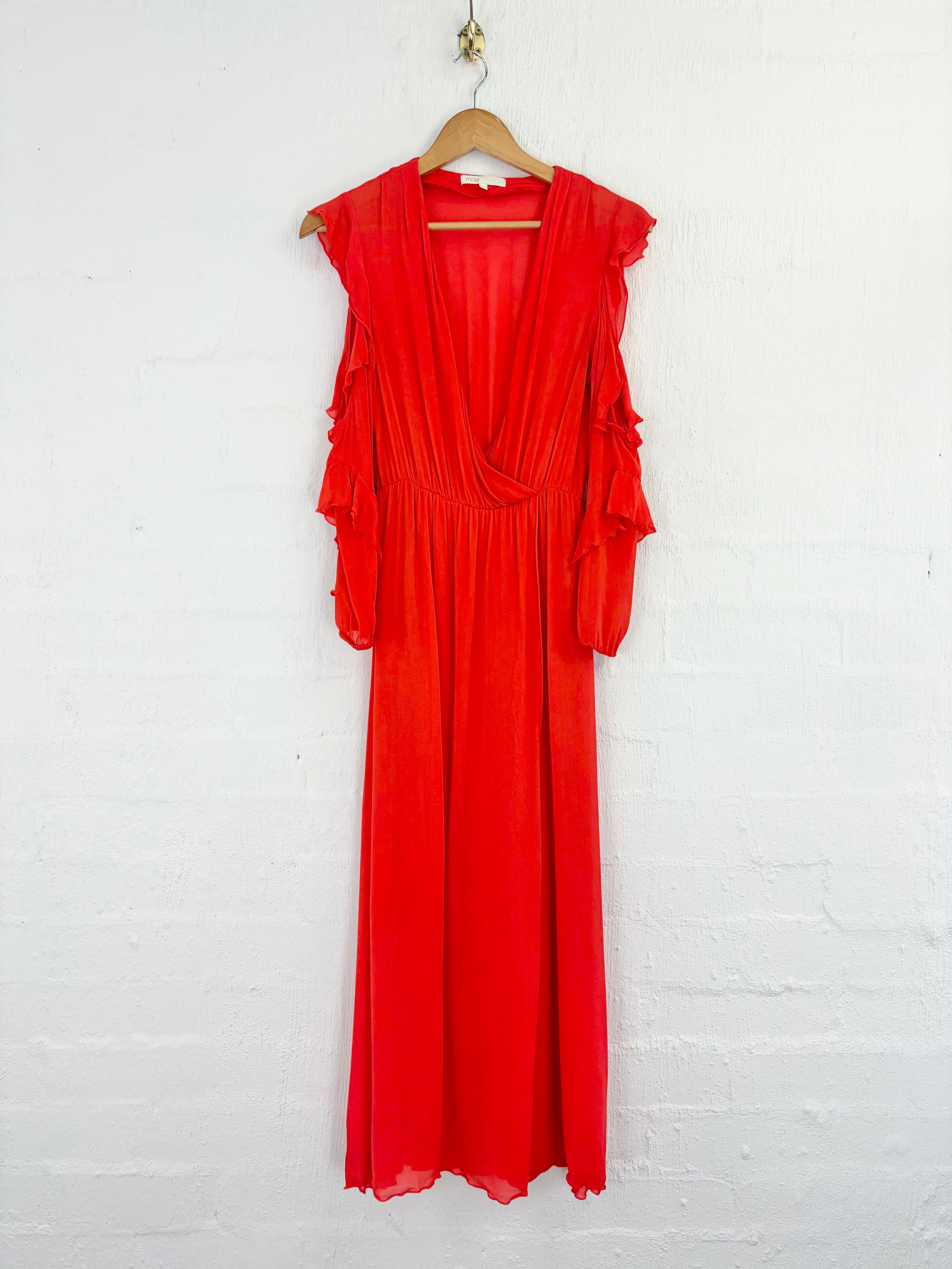 A bright red maxi dress with ruffled cap sleeves and a deep V-neckline, hanging on a wooden hanger against a white painted brick wall.