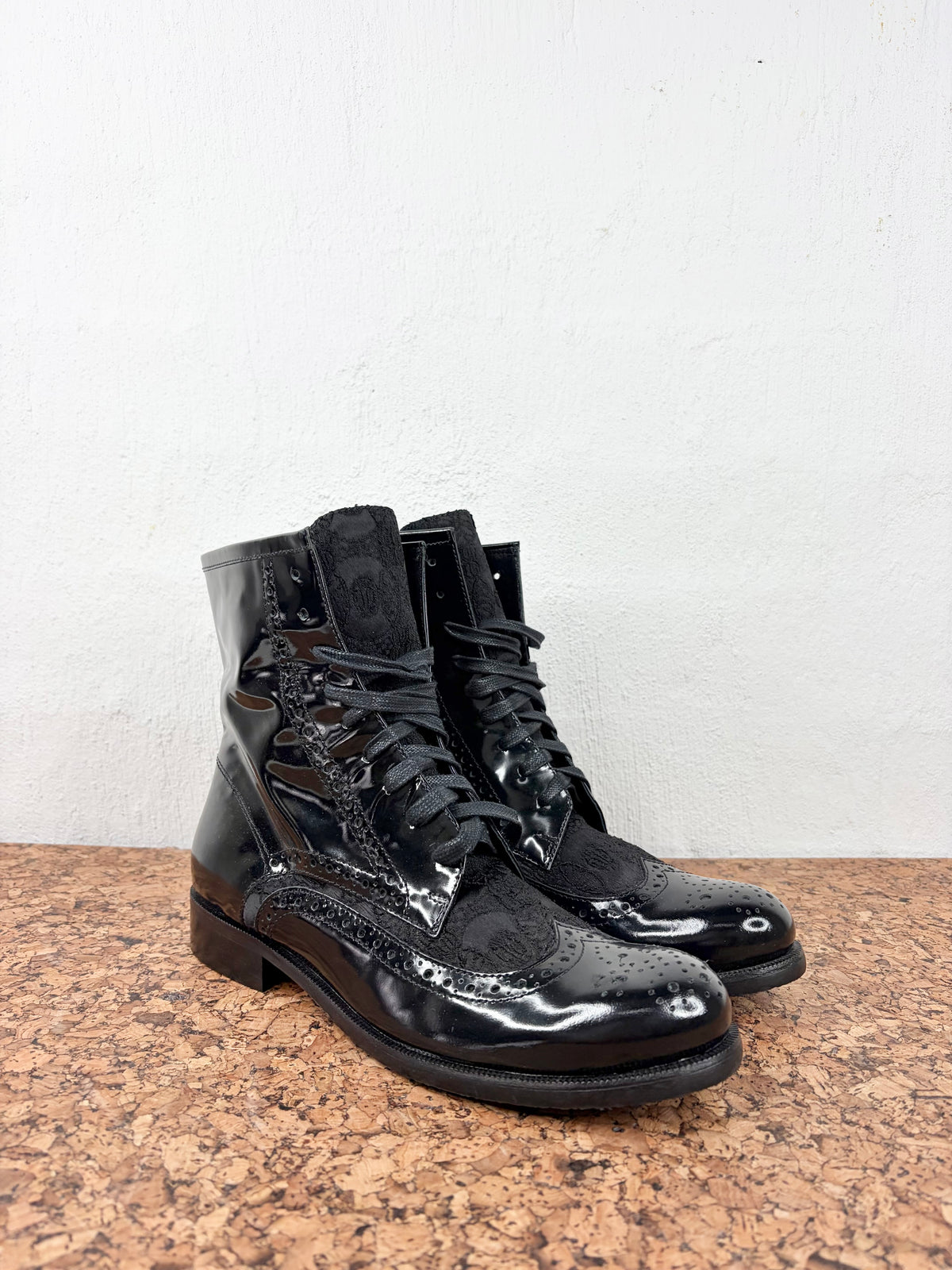 A pair of black patent leather lace-up boots with brogue detailing, displayed on a cork surface against a white wall.