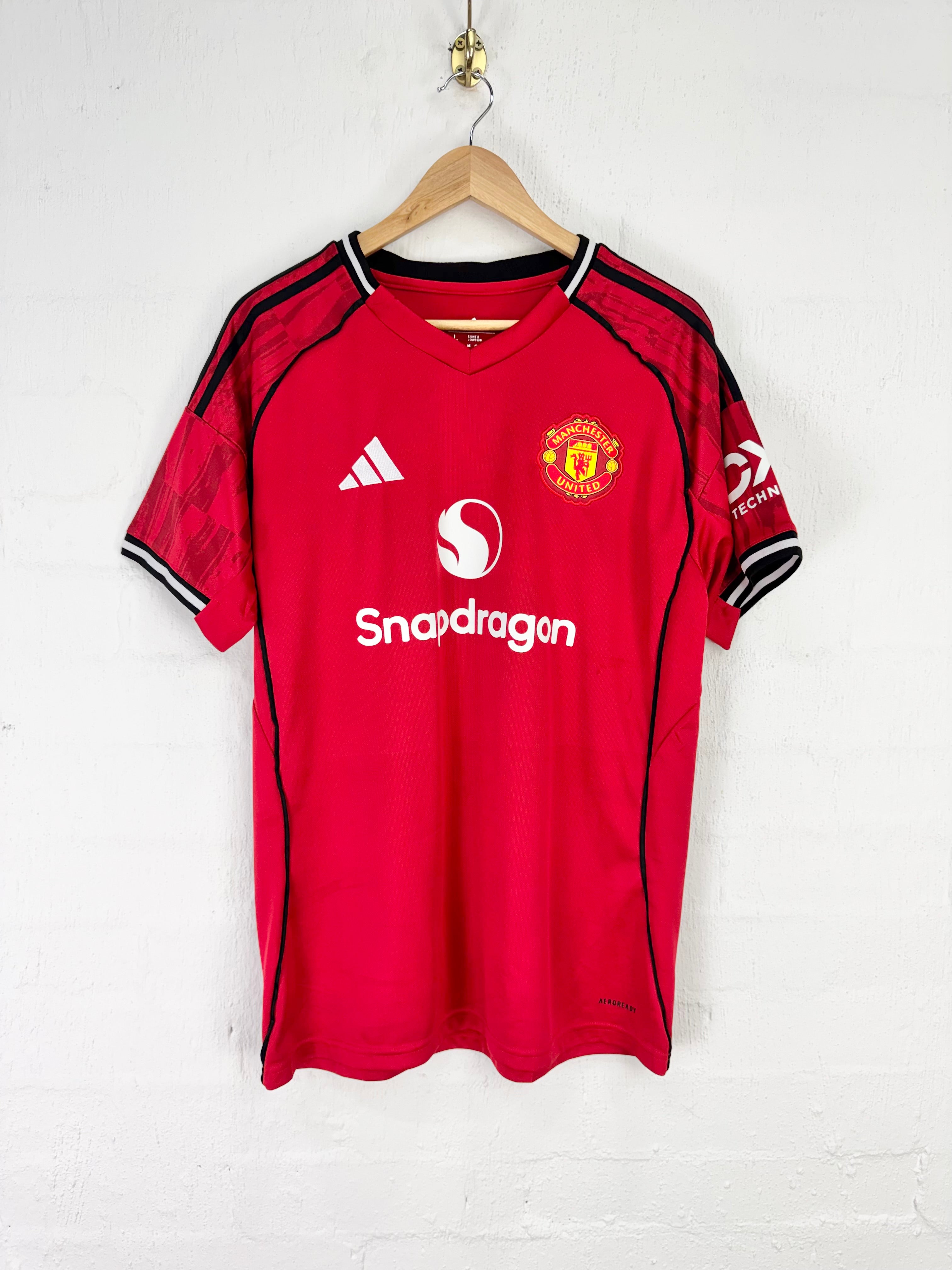 A red Manchester United soccer jersey by Adidas, featuring the Snapdragon sponsor logo, hanging on a wooden hanger against a white brick wall.