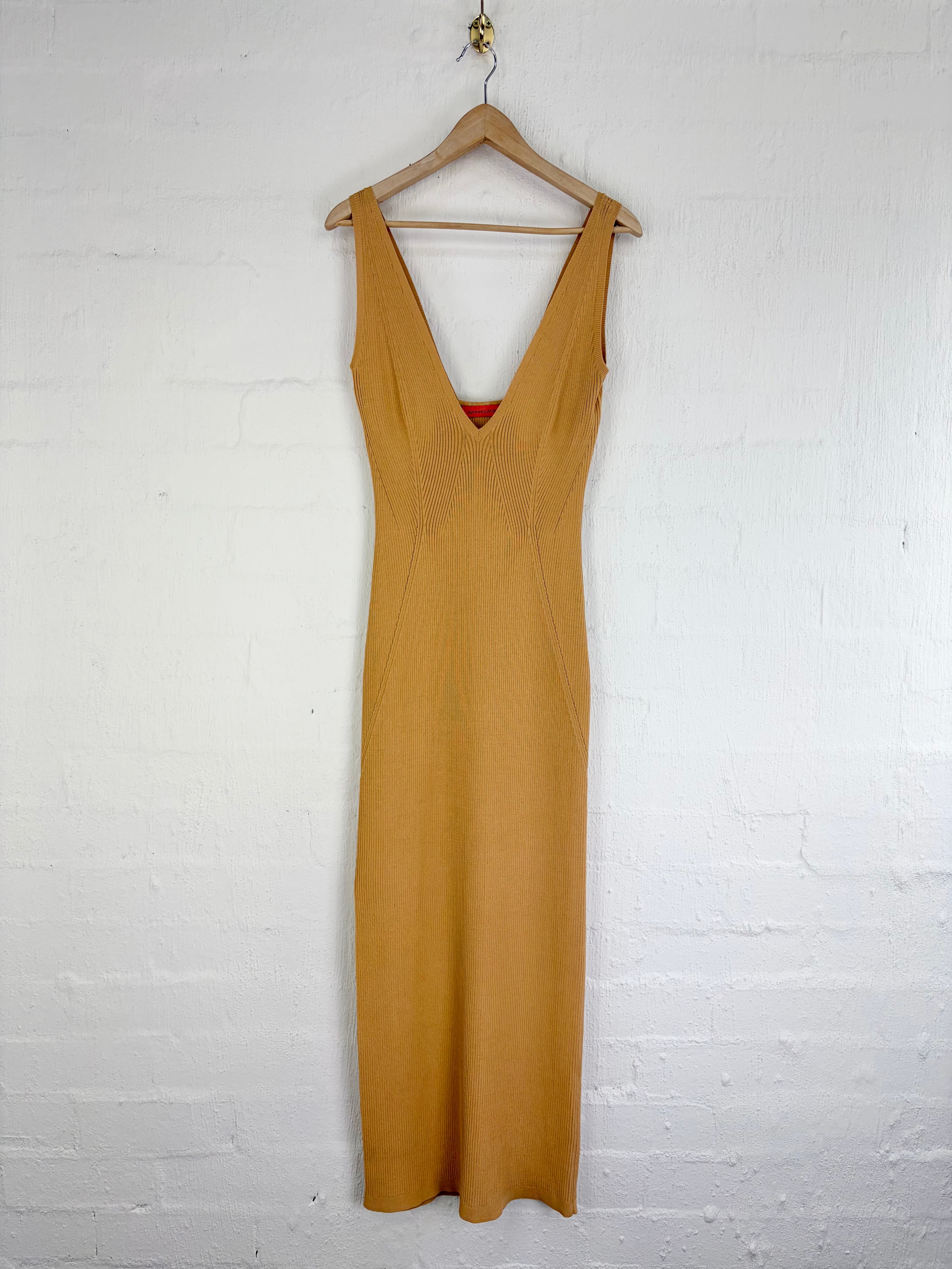 A sleeveless mustard yellow midi dress with a deep V-neckline, hanging on a wooden hanger against a white painted brick wall.