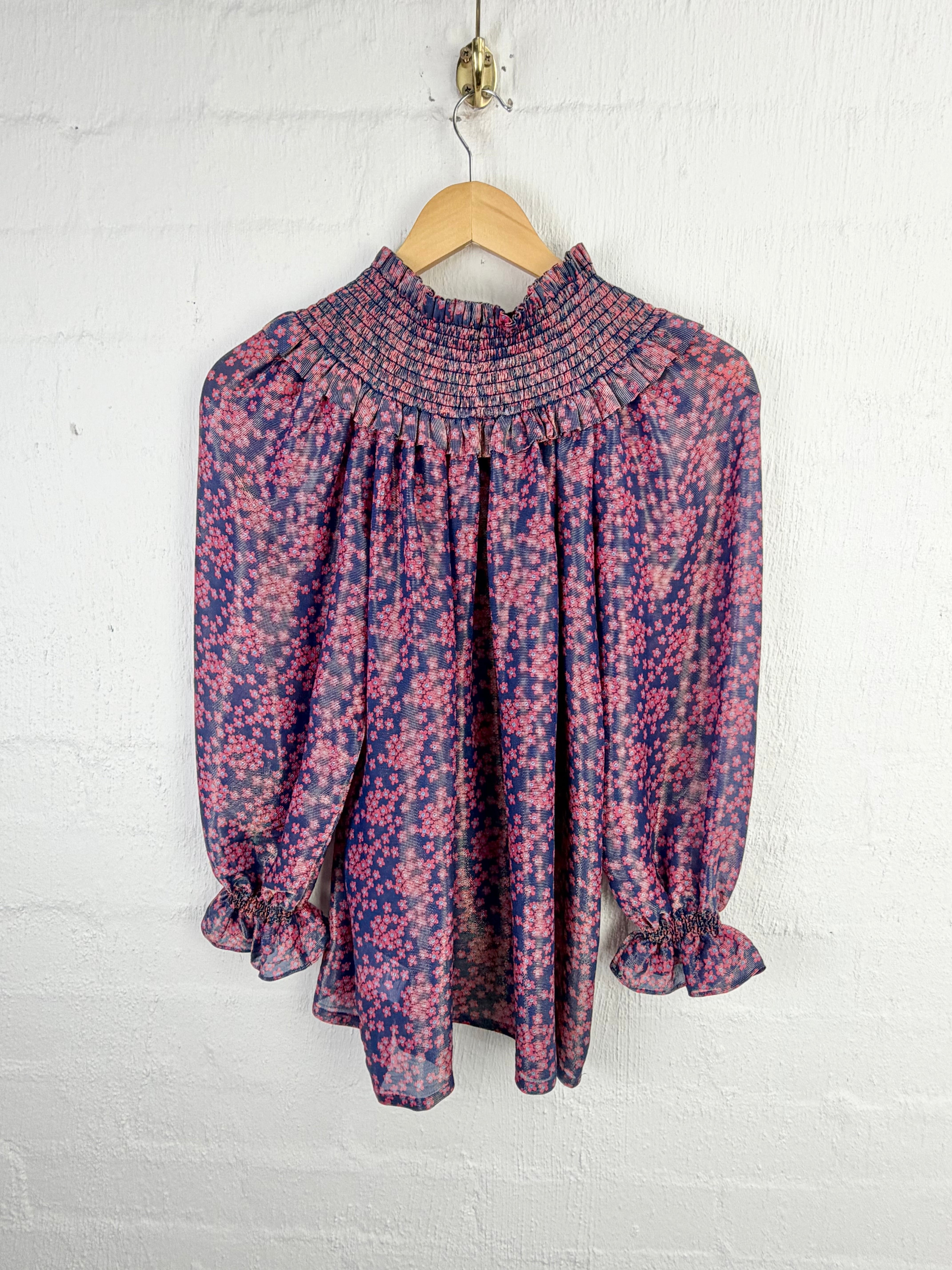 A long-sleeve blouse with a smocked high neckline and ruffled cuffs, featuring a pink and blue floral pattern, hanging on a wooden hanger against a white painted brick wall.