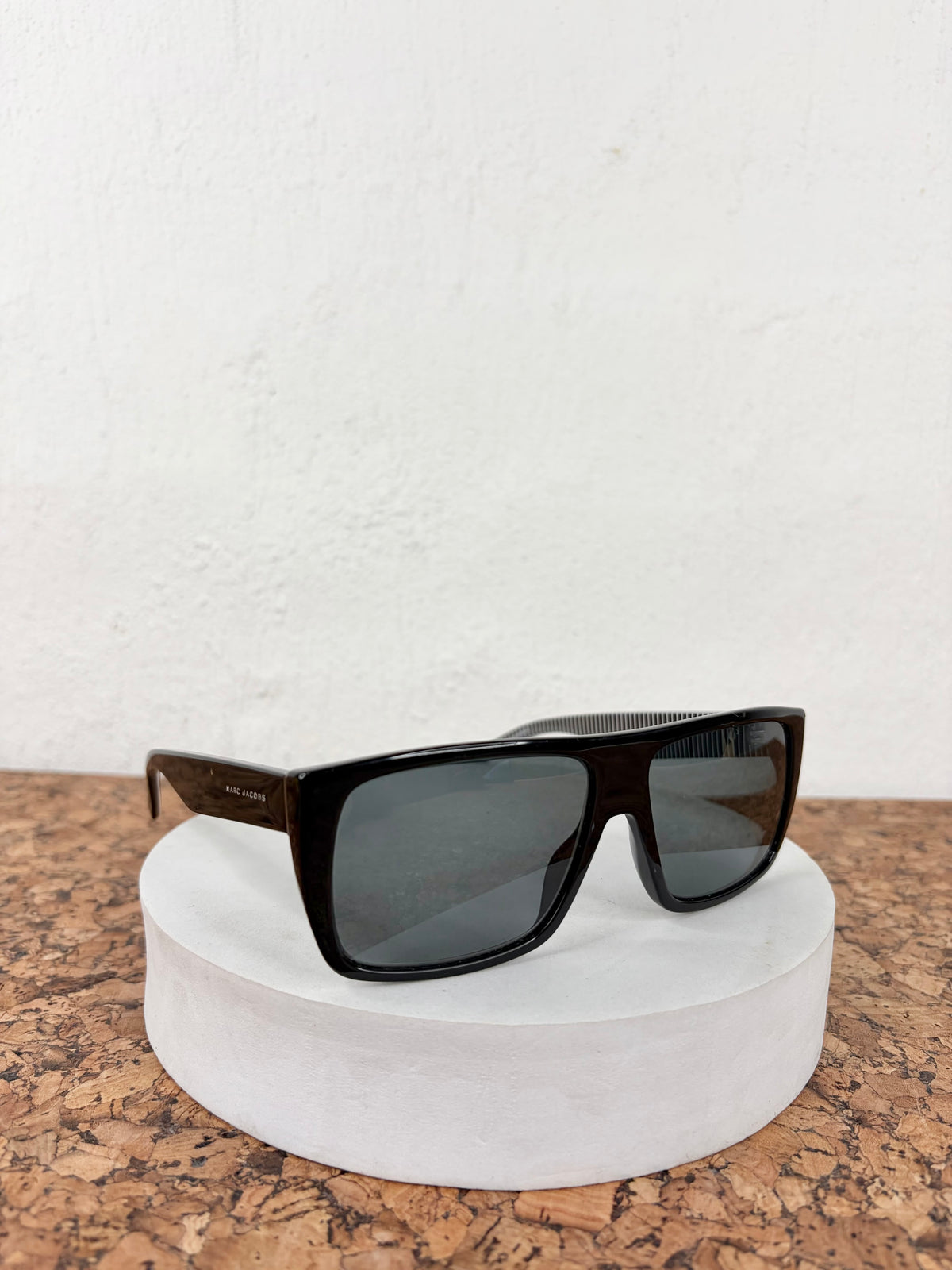 Black square sunglasses with dark lenses displayed on a round white pedestal against a white wall and cork surface.
