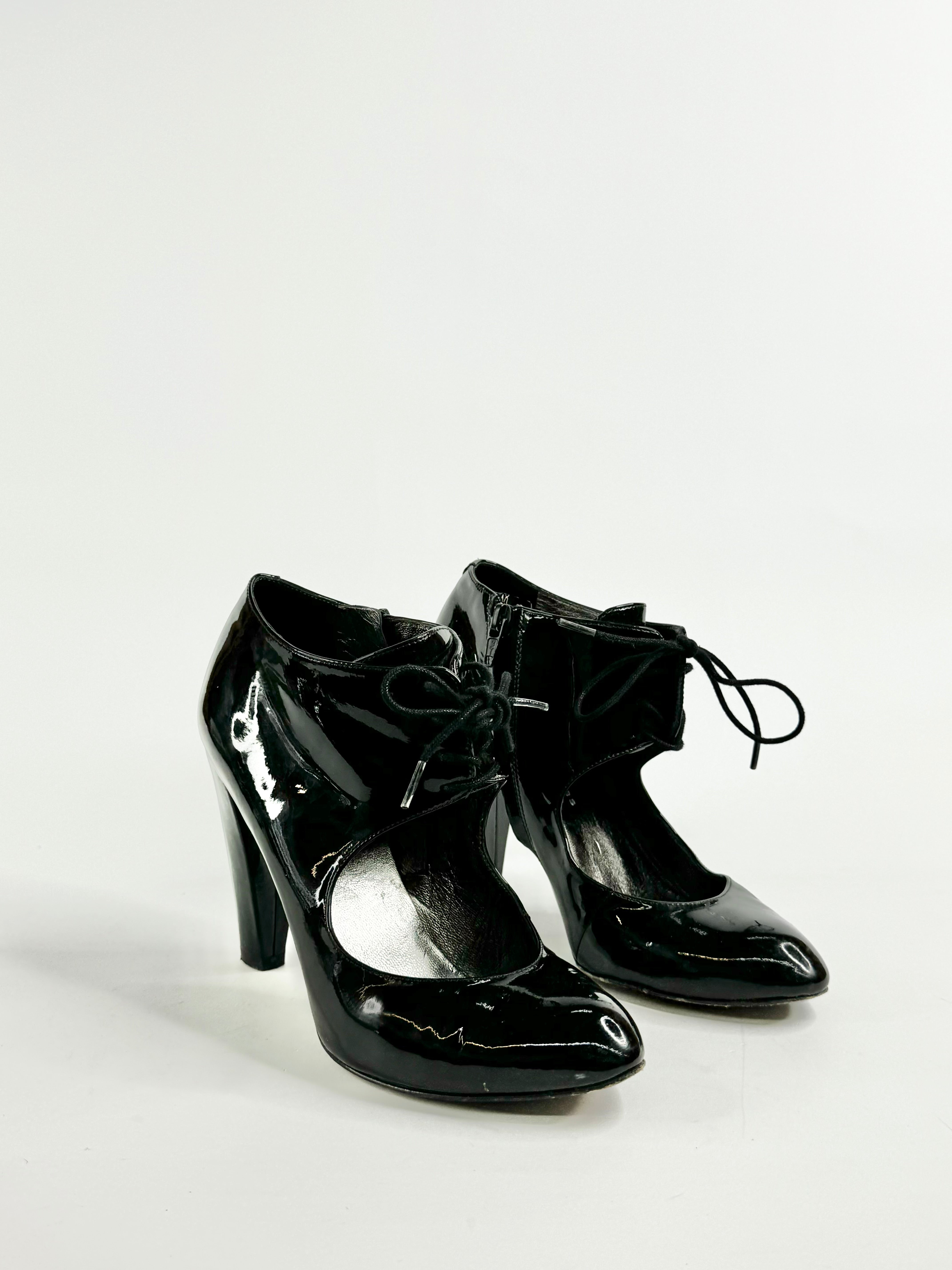 A pair of black patent leather pumps with a lace-up front and a side zipper, on a white background.