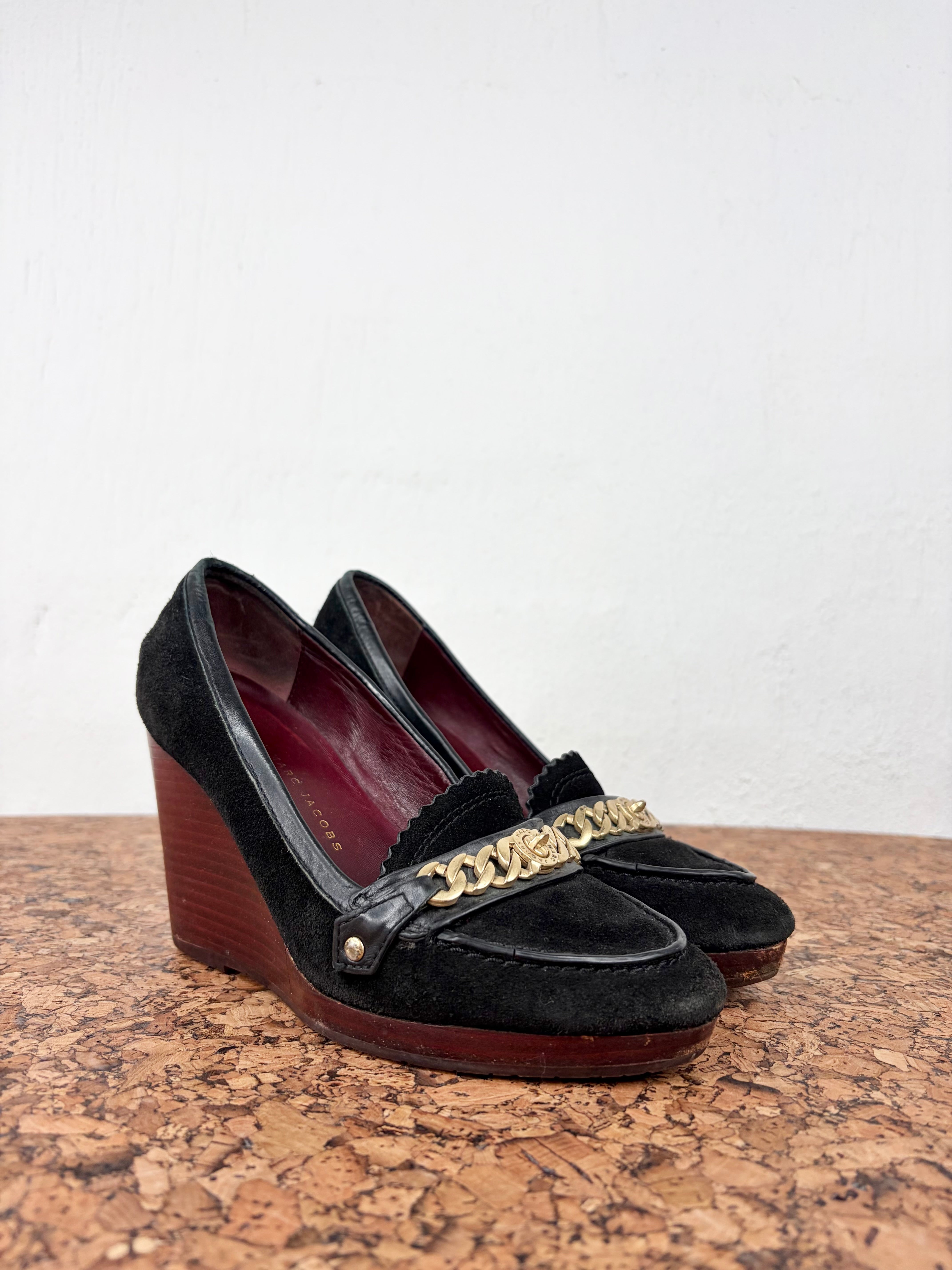 A pair of black suede wedge pumps with gold-tone chain and buckle strap details, displayed on a cork surface against a white wall.