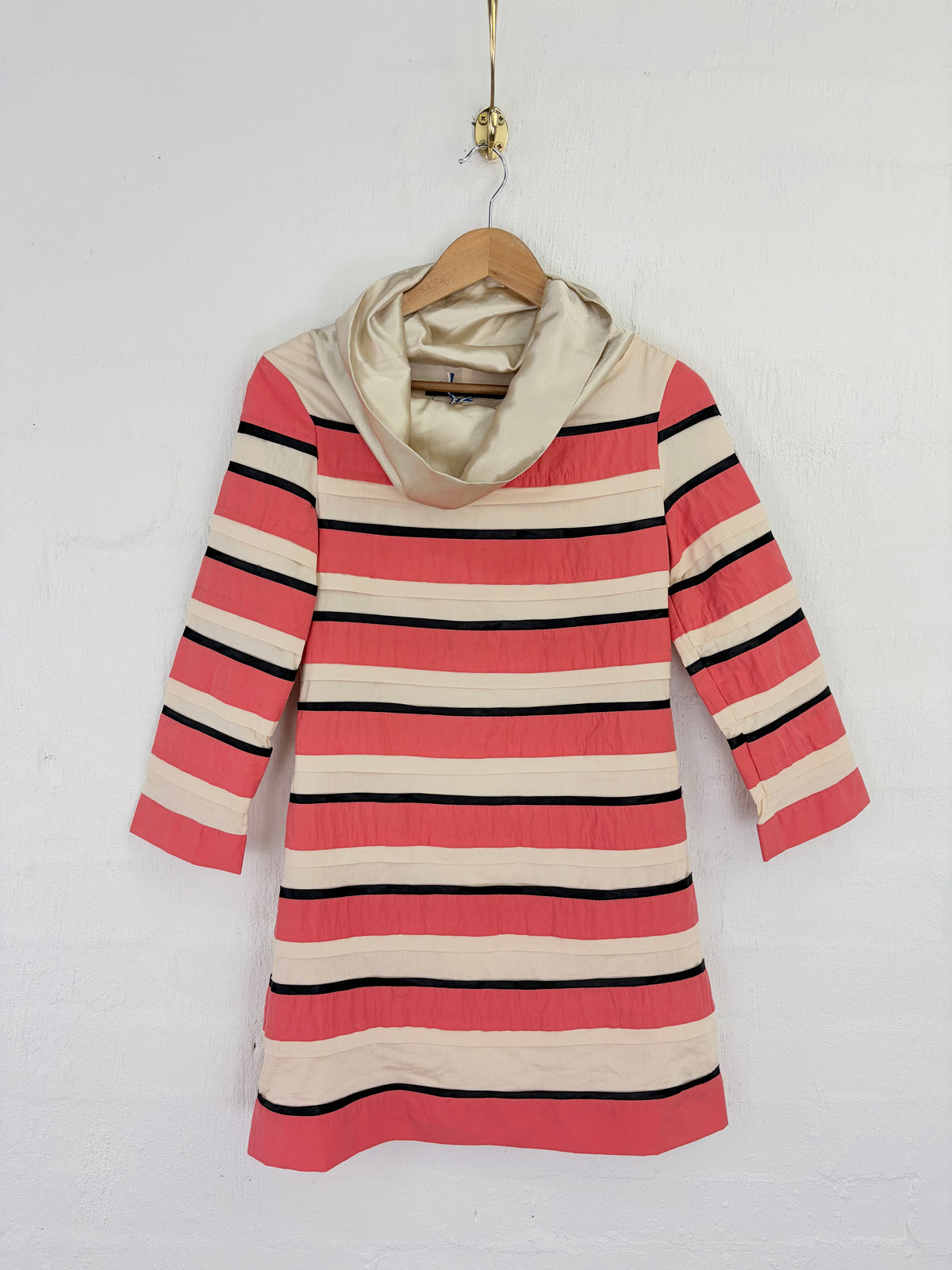 A striped long-sleeve tunic dress with a cowl neck, featuring horizontal stripes of coral pink, cream, and black, hanging on a wooden hanger against a white painted brick wall.