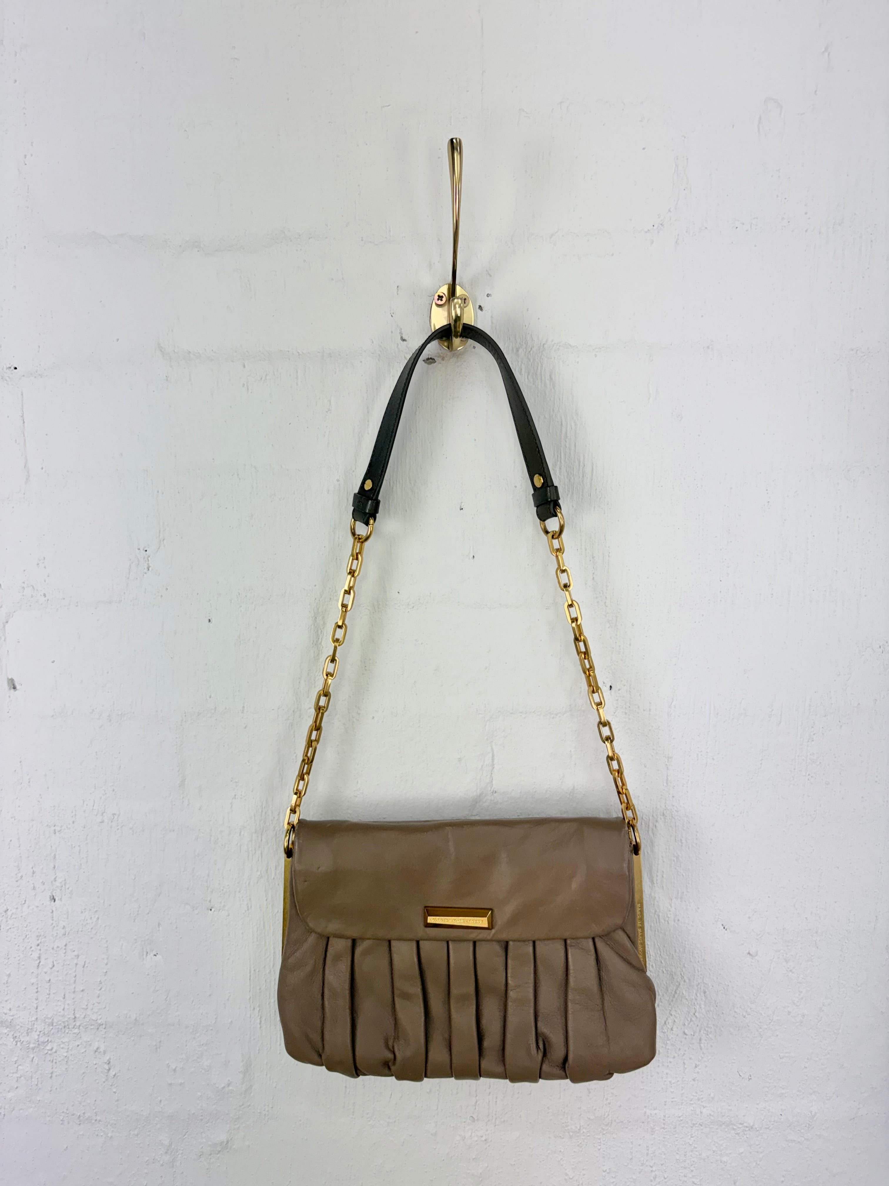 A taupe brown leather shoulder bag with pleated detailing and a black strap featuring gold-tone chain links, hanging from a gold hook against a white brick wall.