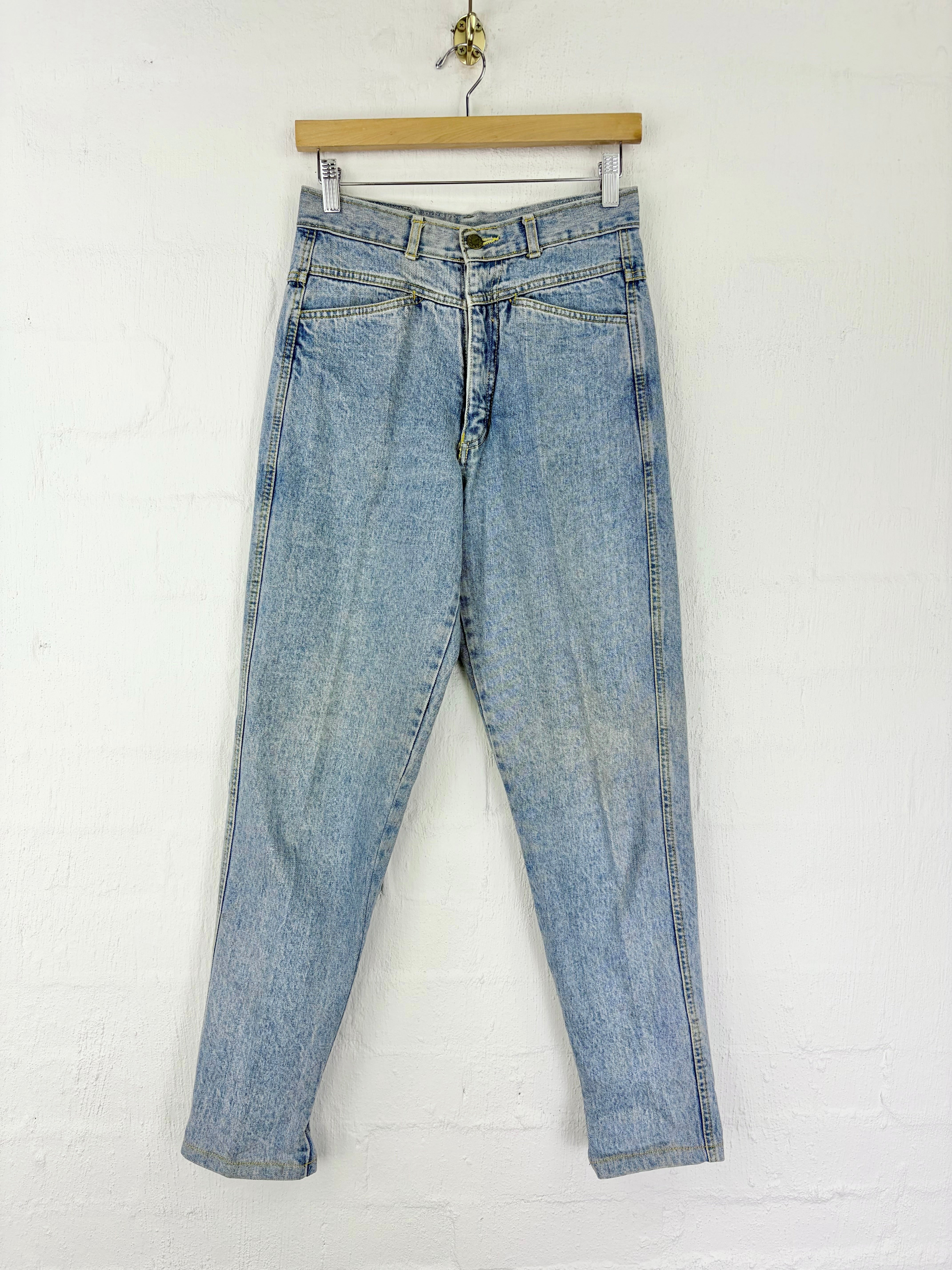 Light wash blue denim jeans hanging from a wooden hanger against a white painted brick wall.