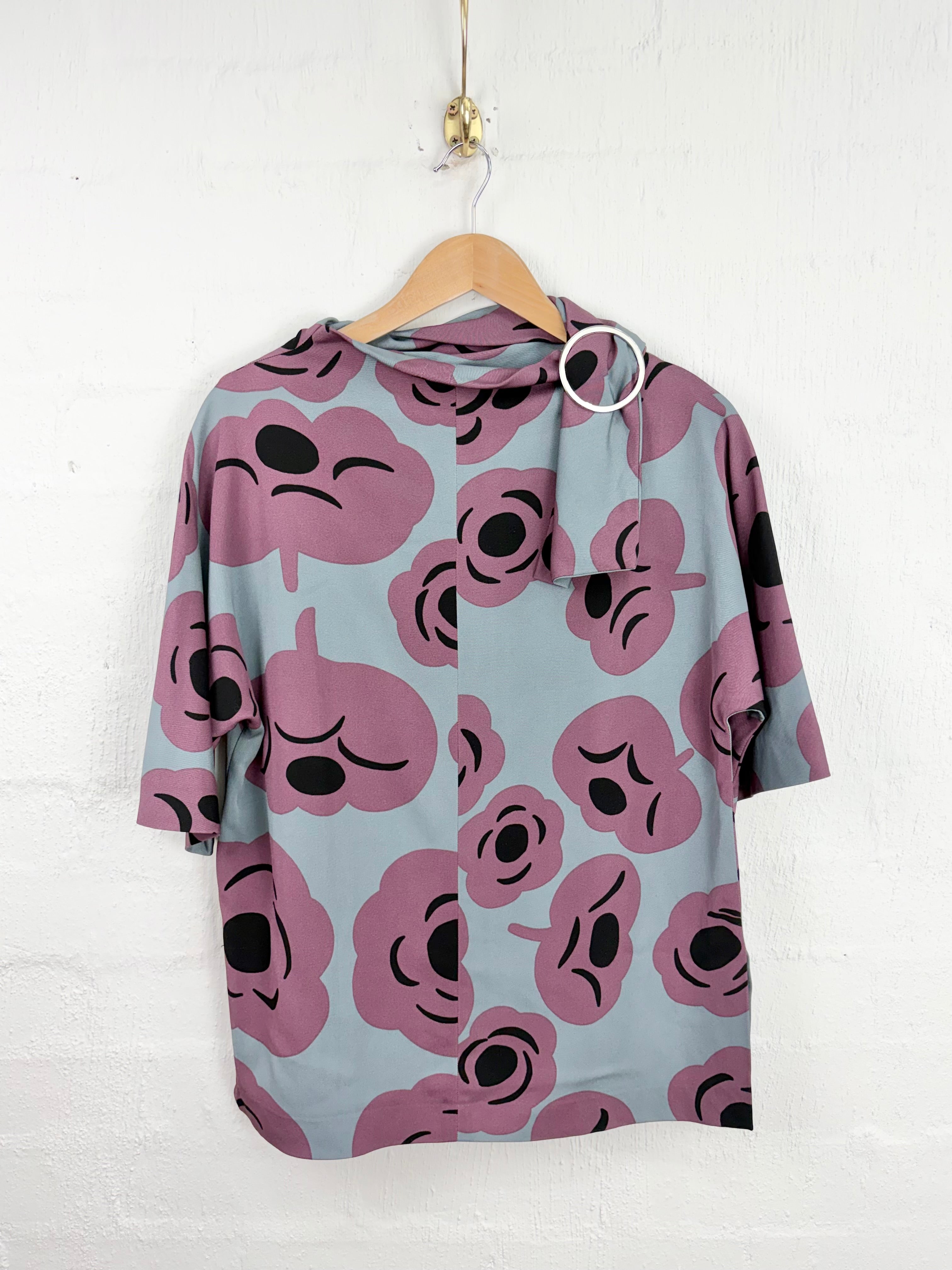 A short-sleeve button-up shirt with a light gray base and a pink and black cartoon face pattern, hanging on a wooden hanger against a white brick wall.