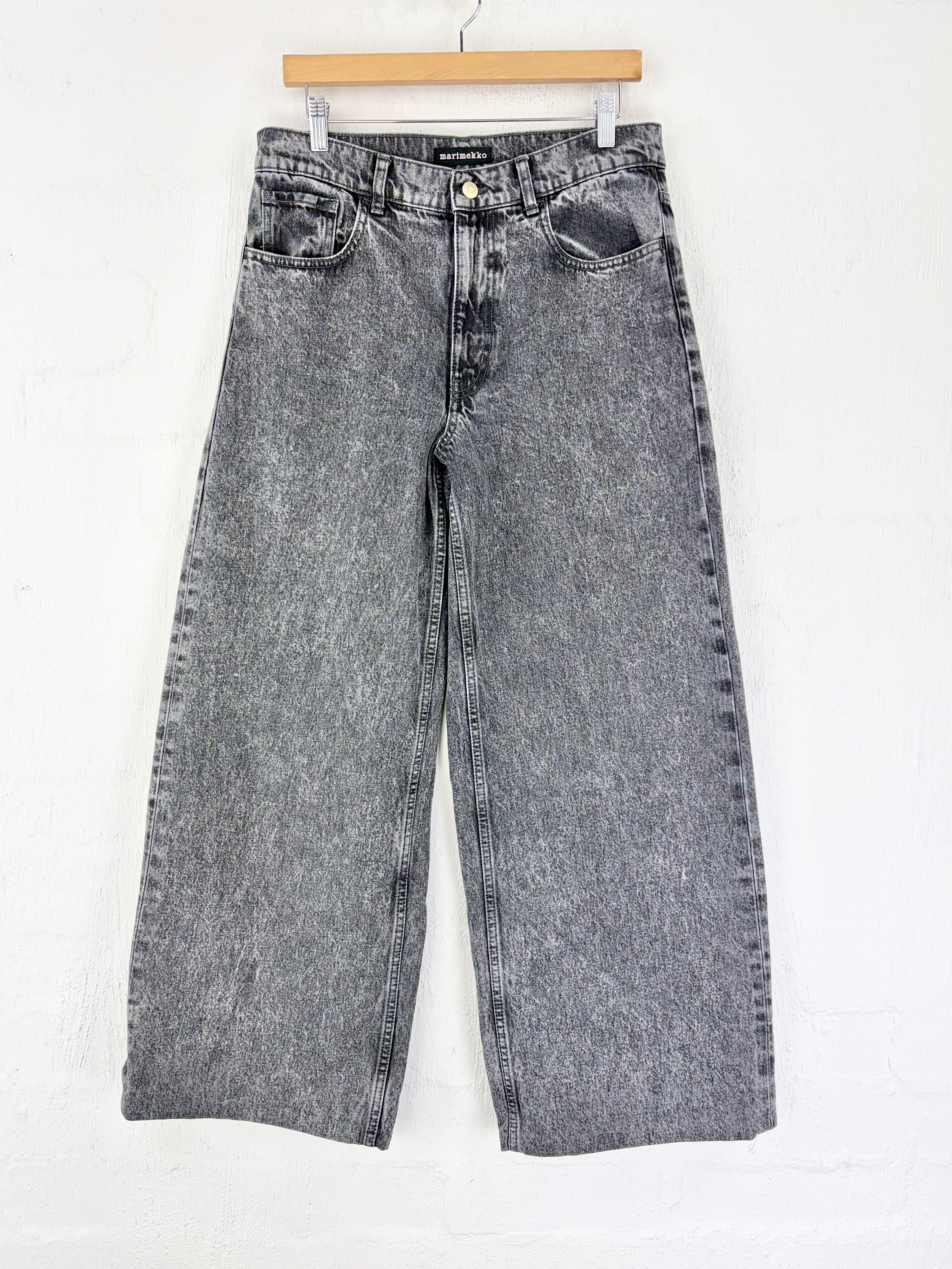 A pair of faded gray wide-leg denim jeans hanging from a wooden hanger against a white textured wall.