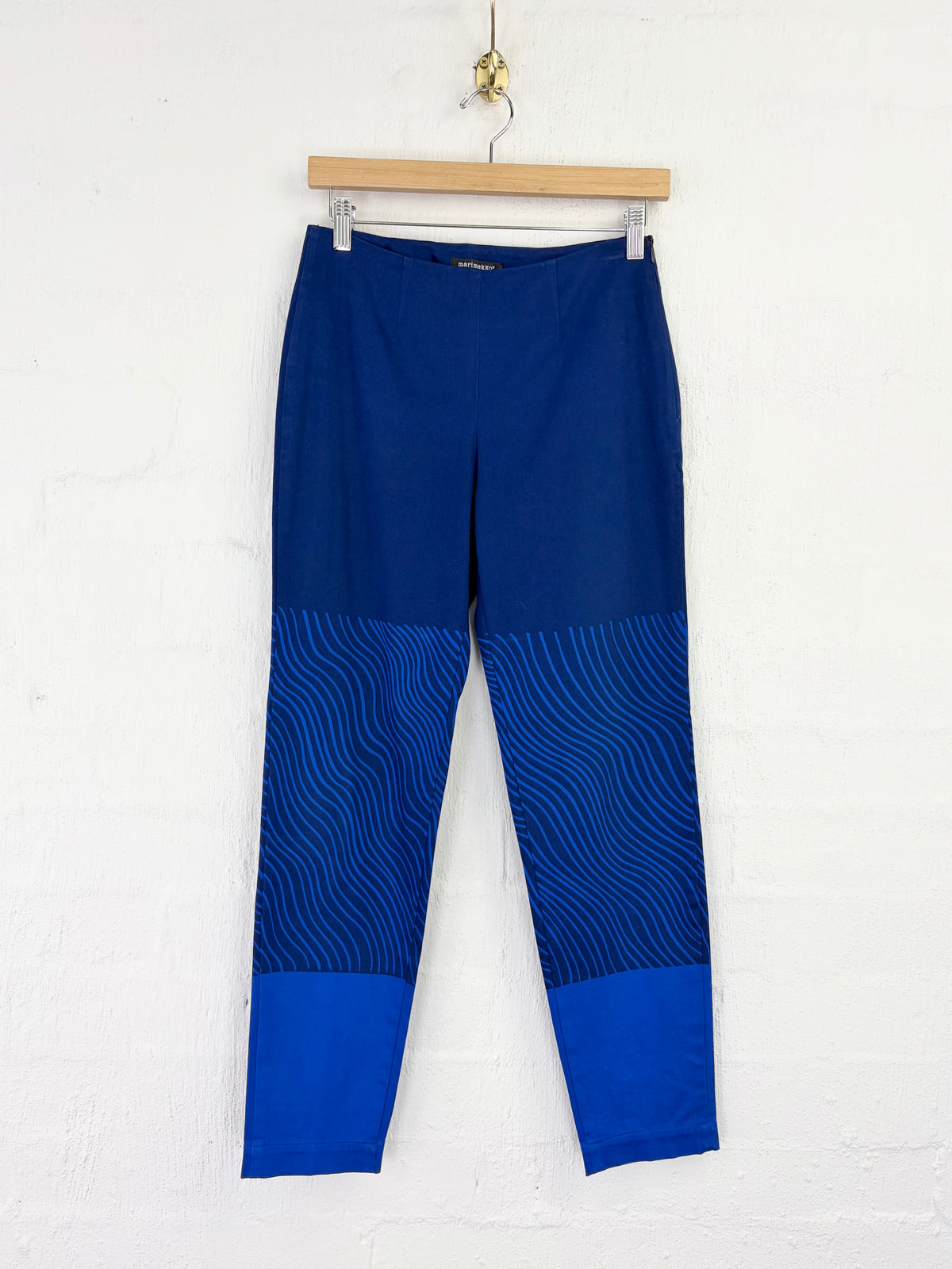 Blue cropped pants with a wavy patterned lower leg and solid blue cuffs, hanging from a wooden hanger against a white wall.