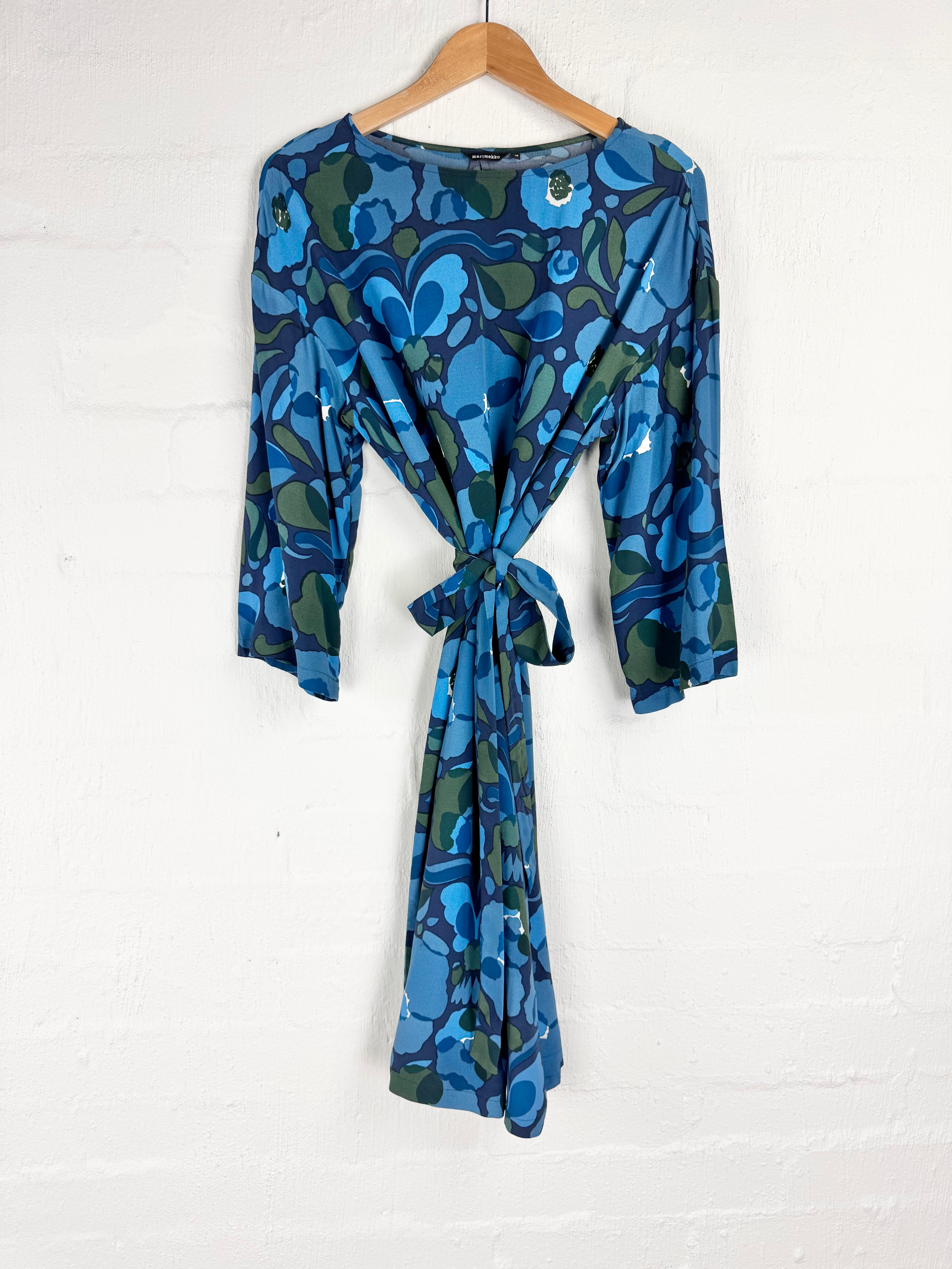 A blue and green abstract floral print wrap dress with three-quarter sleeves, hanging on a wooden hanger against a white painted brick wall.