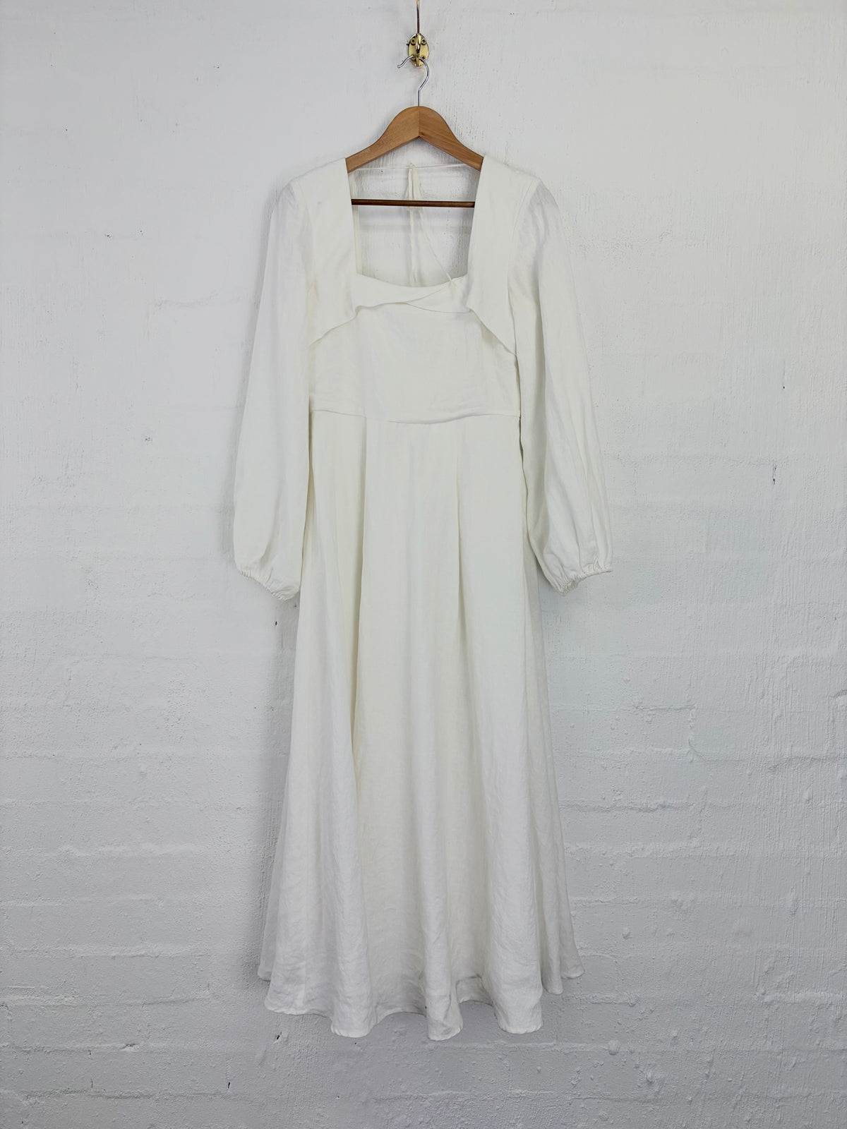 An off-white linen midi dress with three-quarter balloon sleeves and a square neckline, hanging on a wooden hanger against a white painted brick wall.