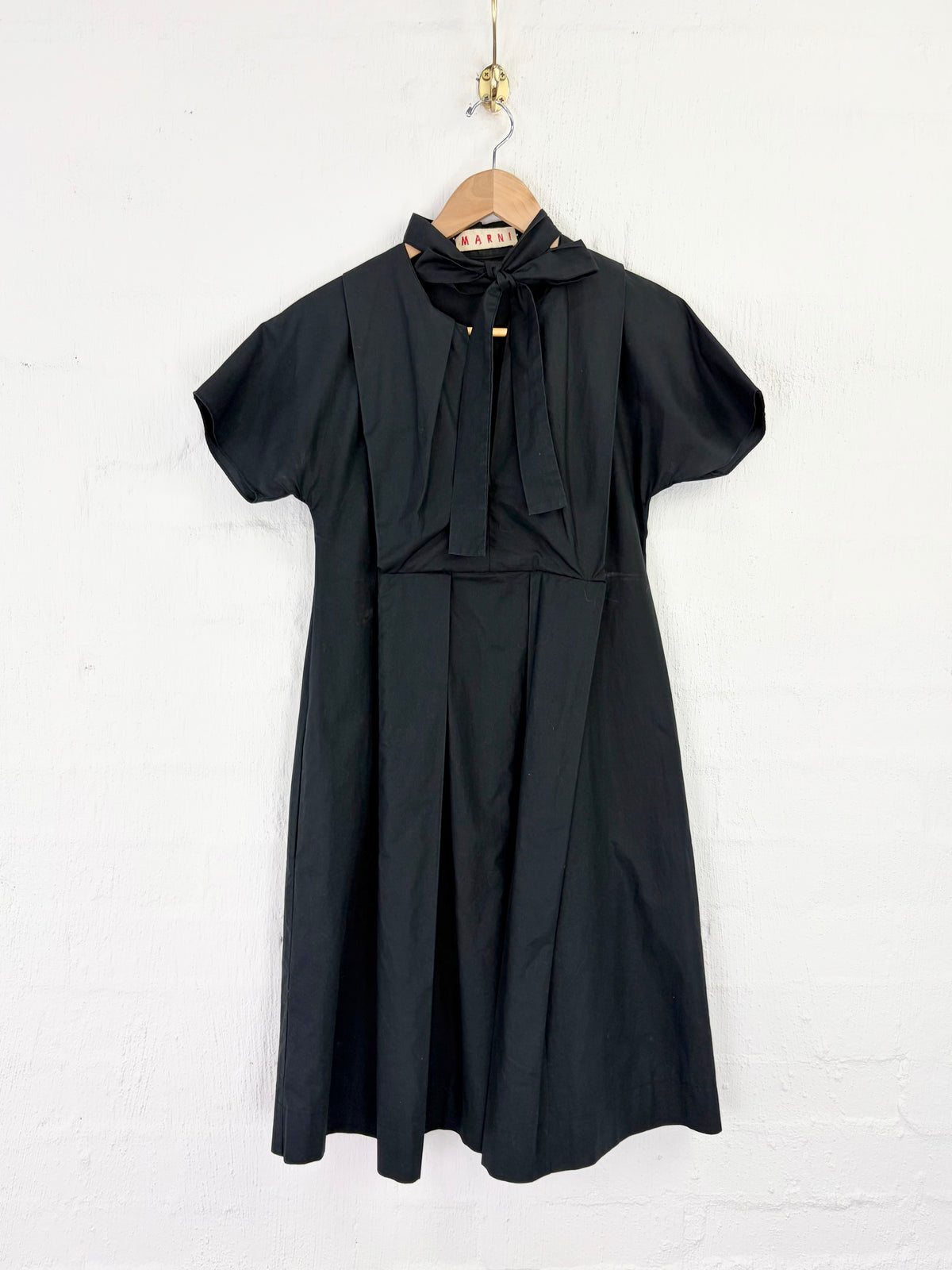 A black short-sleeve dress with a bow tie neckline, hanging on a wooden hanger against a white painted brick wall.