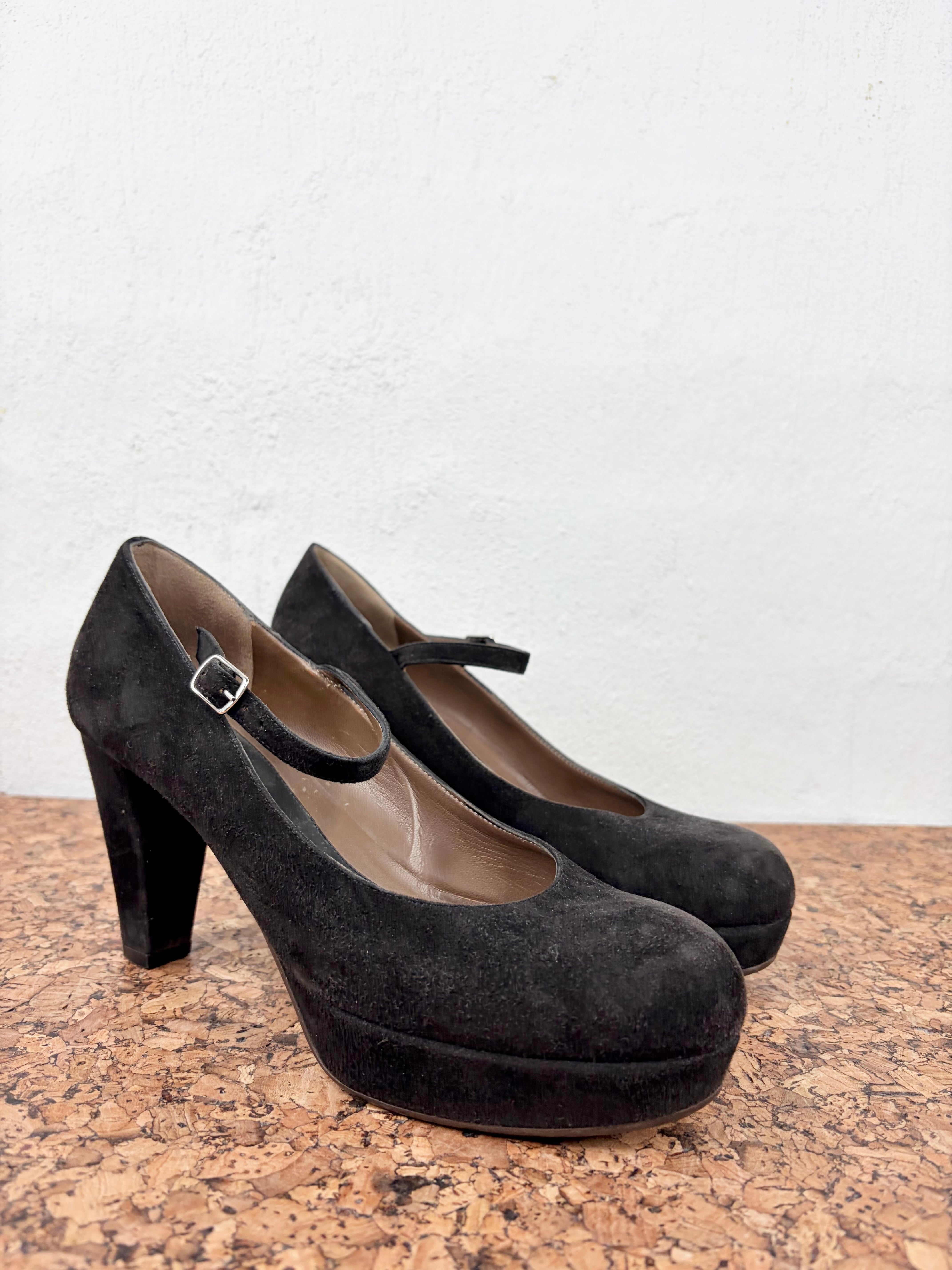 A pair of black suede Mary Jane pumps with a chunky block heel and platform sole, resting on a cork surface against a white wall.