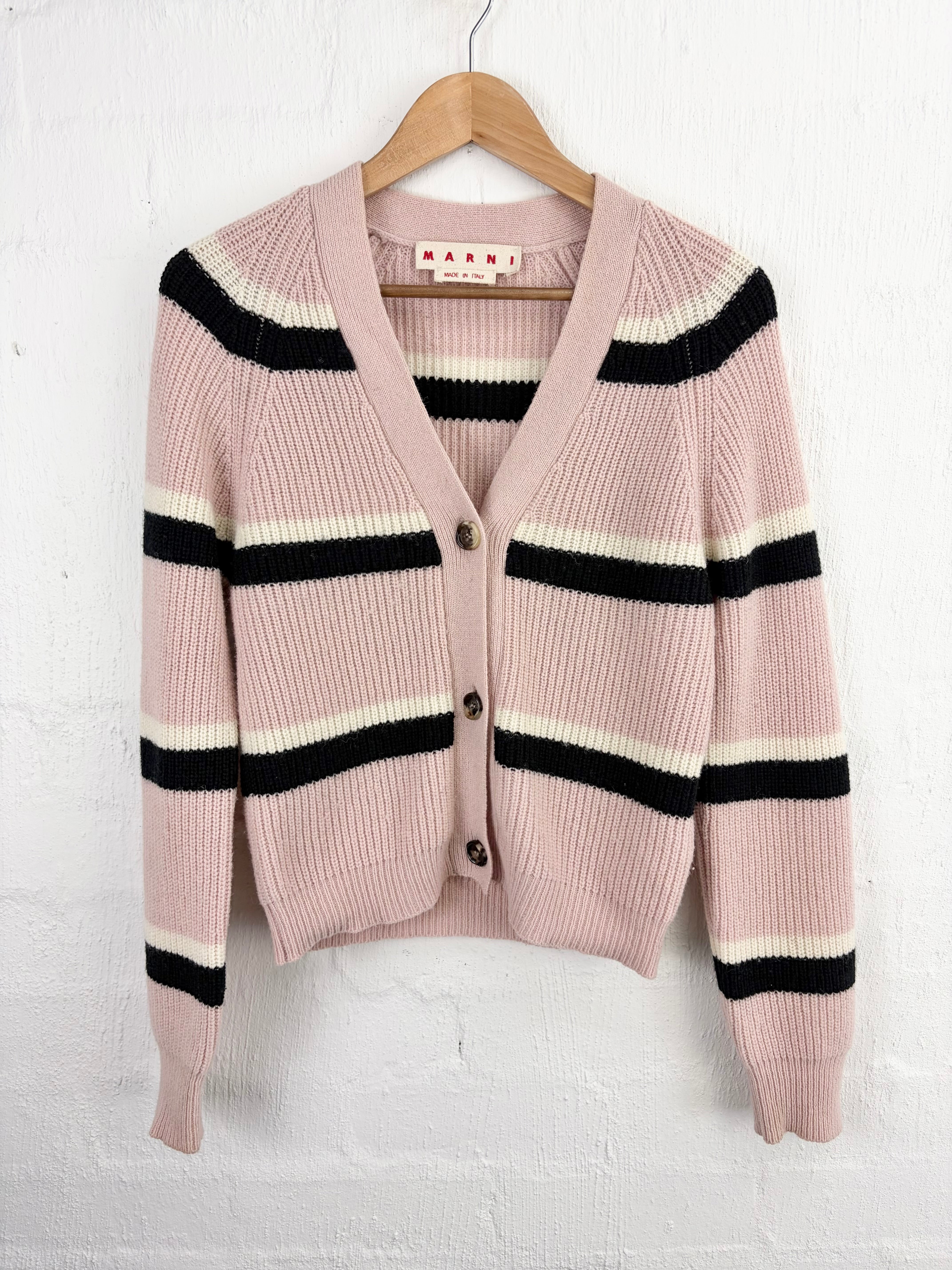 A pink ribbed knit cardigan sweater with horizontal black and cream stripes, hanging on a wooden hanger against a white painted brick wall. The brand name 'SABRI' is visible on the inner neck label.