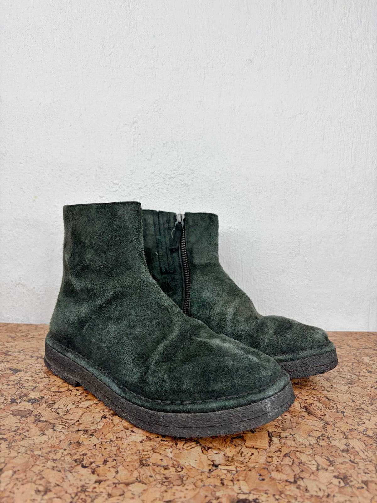 A pair of dark green suede ankle boots with side zippers, resting on a cork surface against a white wall.