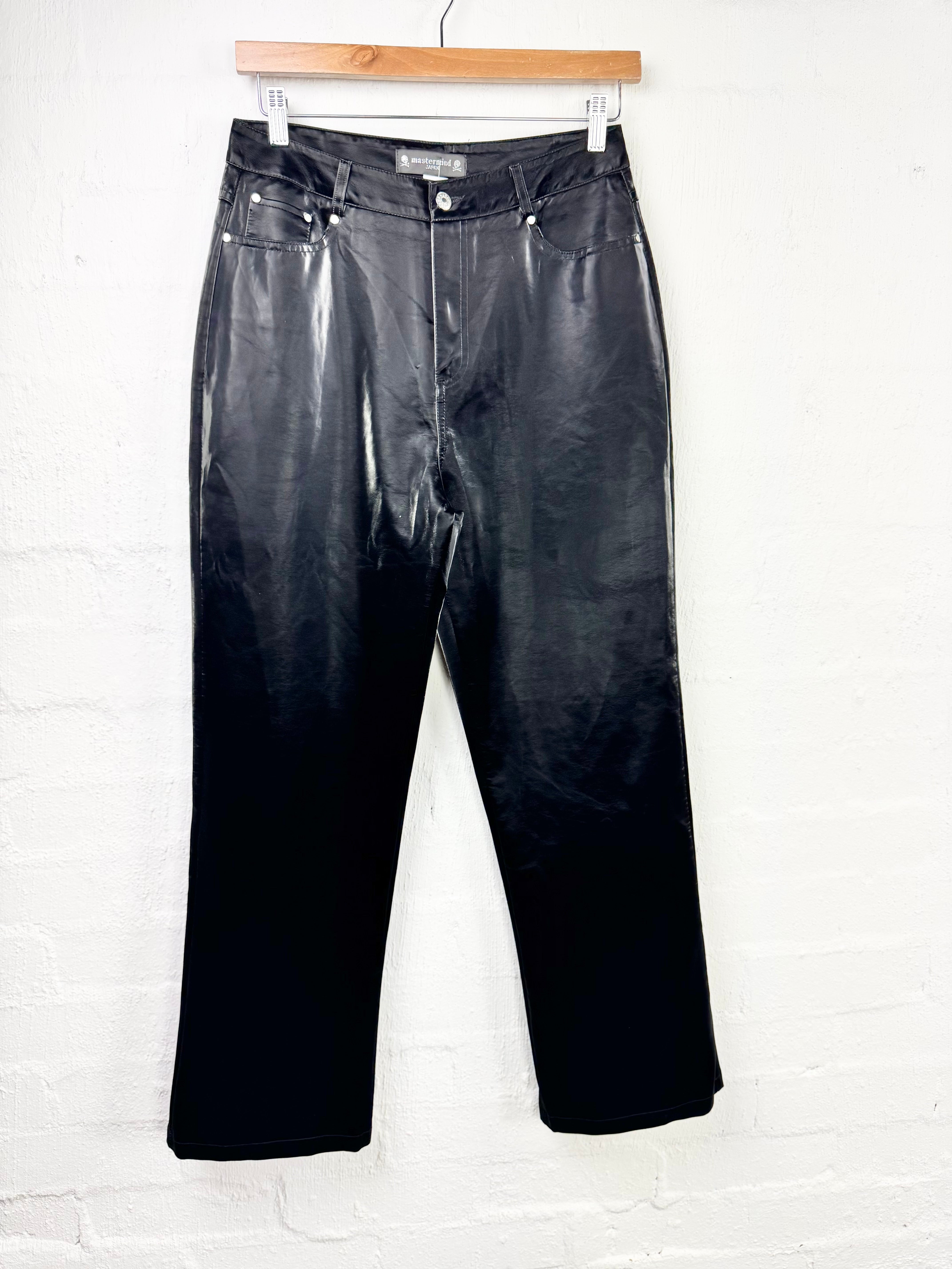 Black faux leather cropped pants hanging from a wooden hanger against a white brick wall.