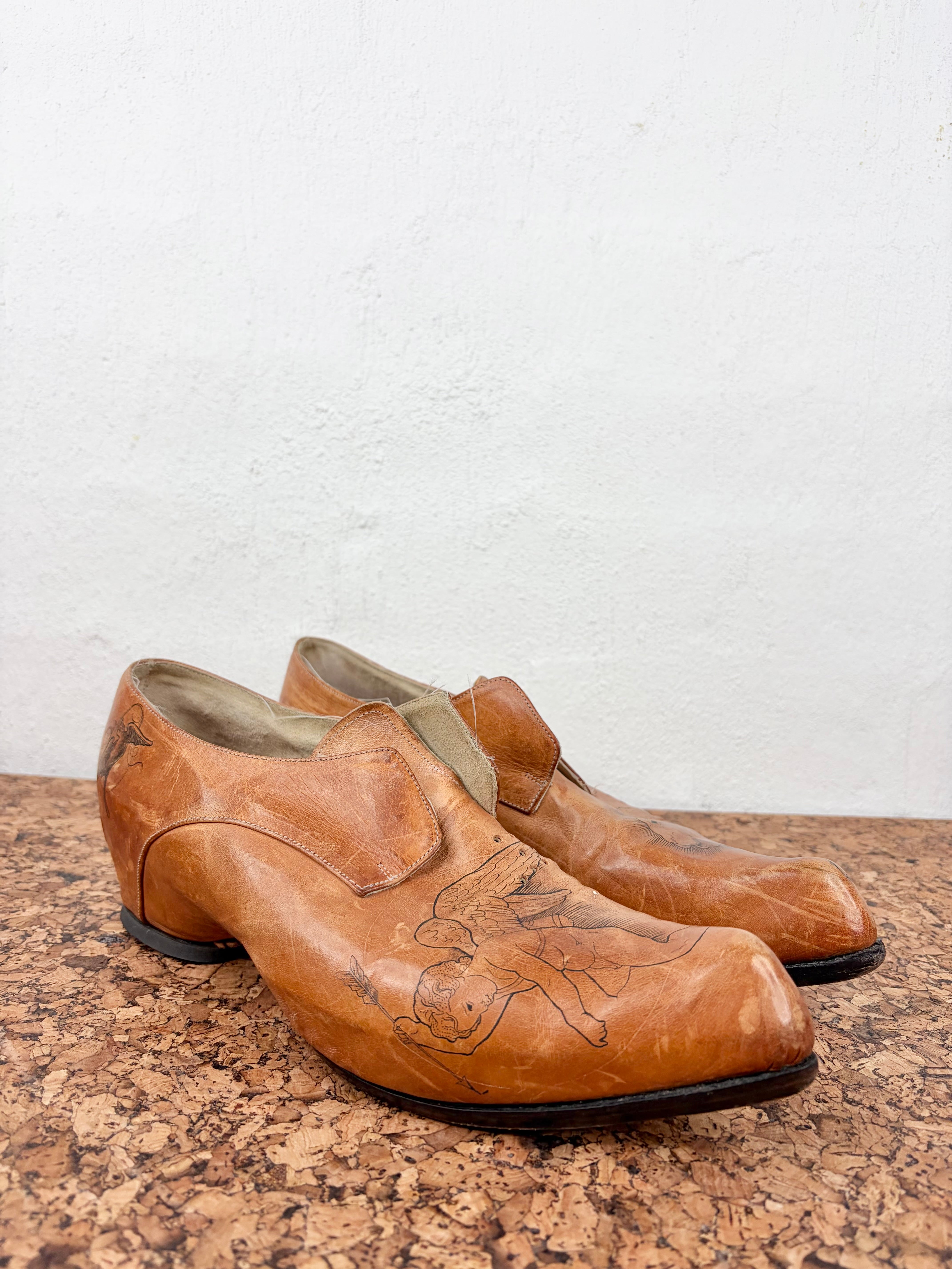 A pair of vintage tan leather slip-on shoes with hand-painted black winged figures on the toe boxes, displayed on a cork surface against a white wall.