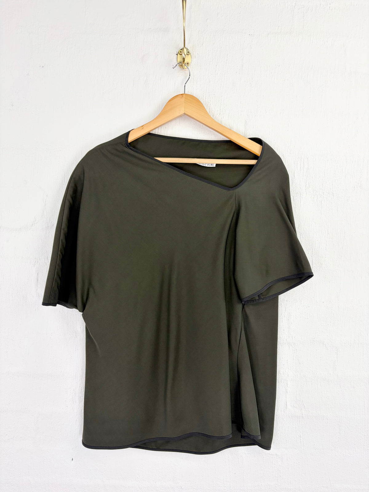 An olive green short-sleeve top with an asymmetrical draped sleeve detail, hanging on a wooden hanger against a white textured wall.