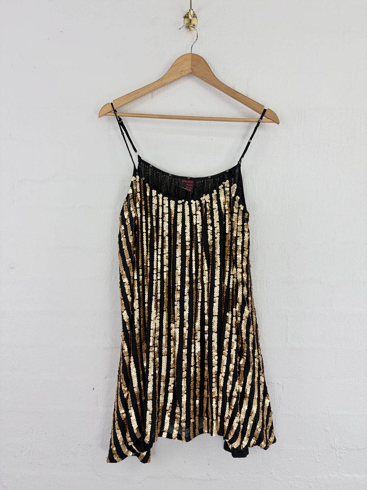 A black and gold vertically striped sequin slip dress hanging on a wooden hanger against a white painted brick wall.