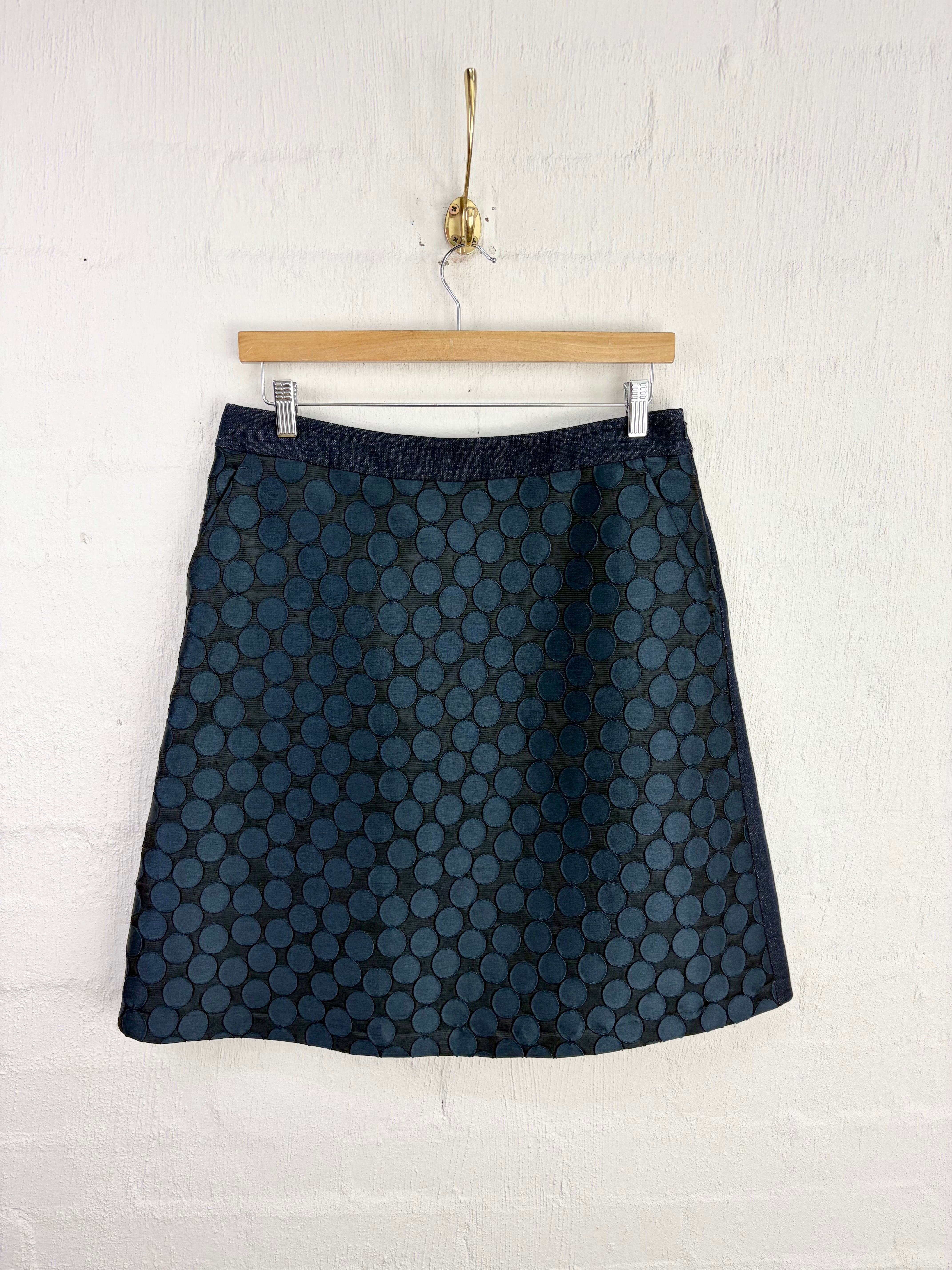 A dark navy blue A-line skirt with a repeating circular geometric pattern, hanging from a wooden hanger against a white painted brick wall.