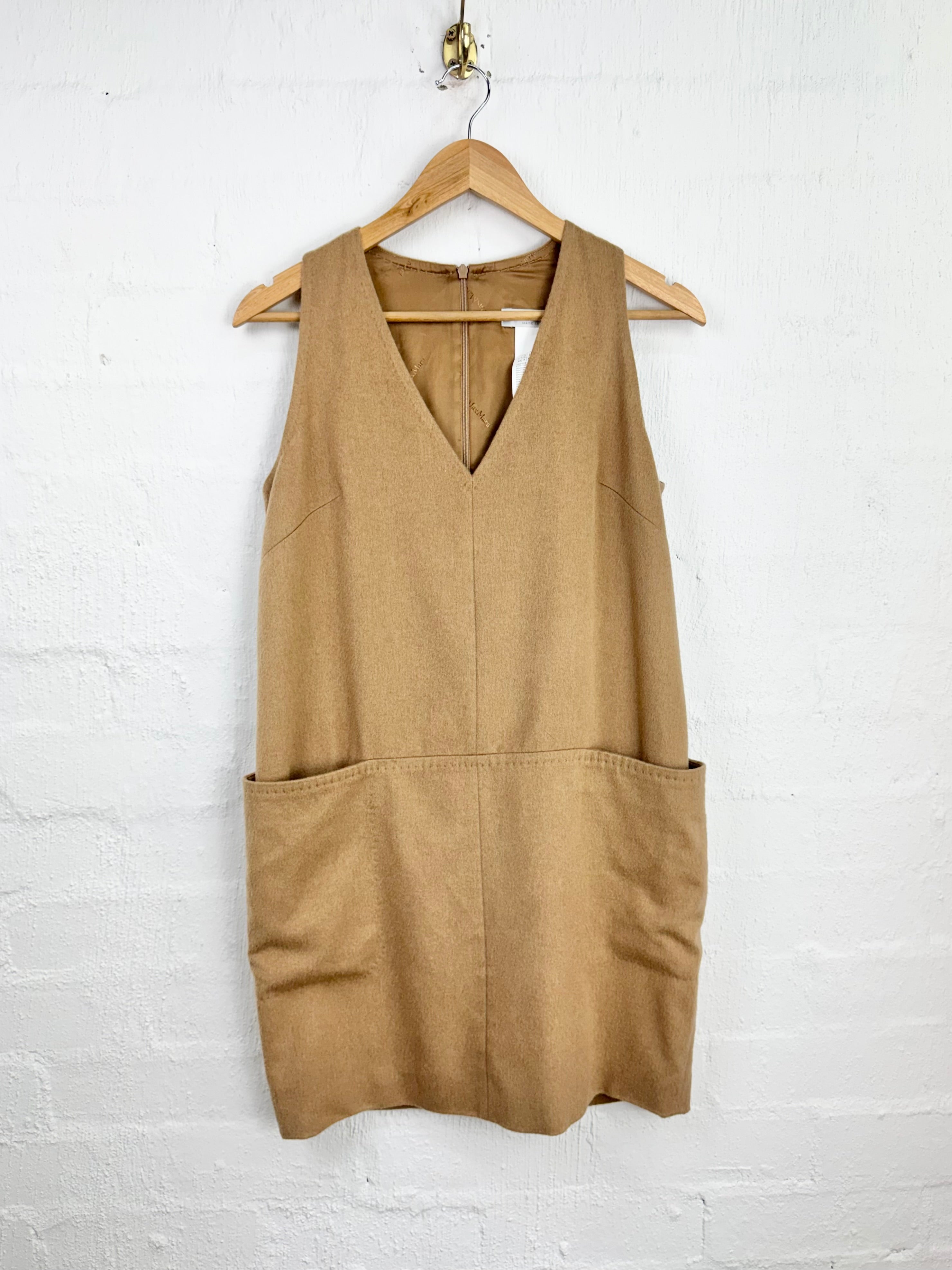 A sleeveless tan utility dress with a V-neckline and large front patch pockets, hanging on a wooden hanger against a white painted brick wall.