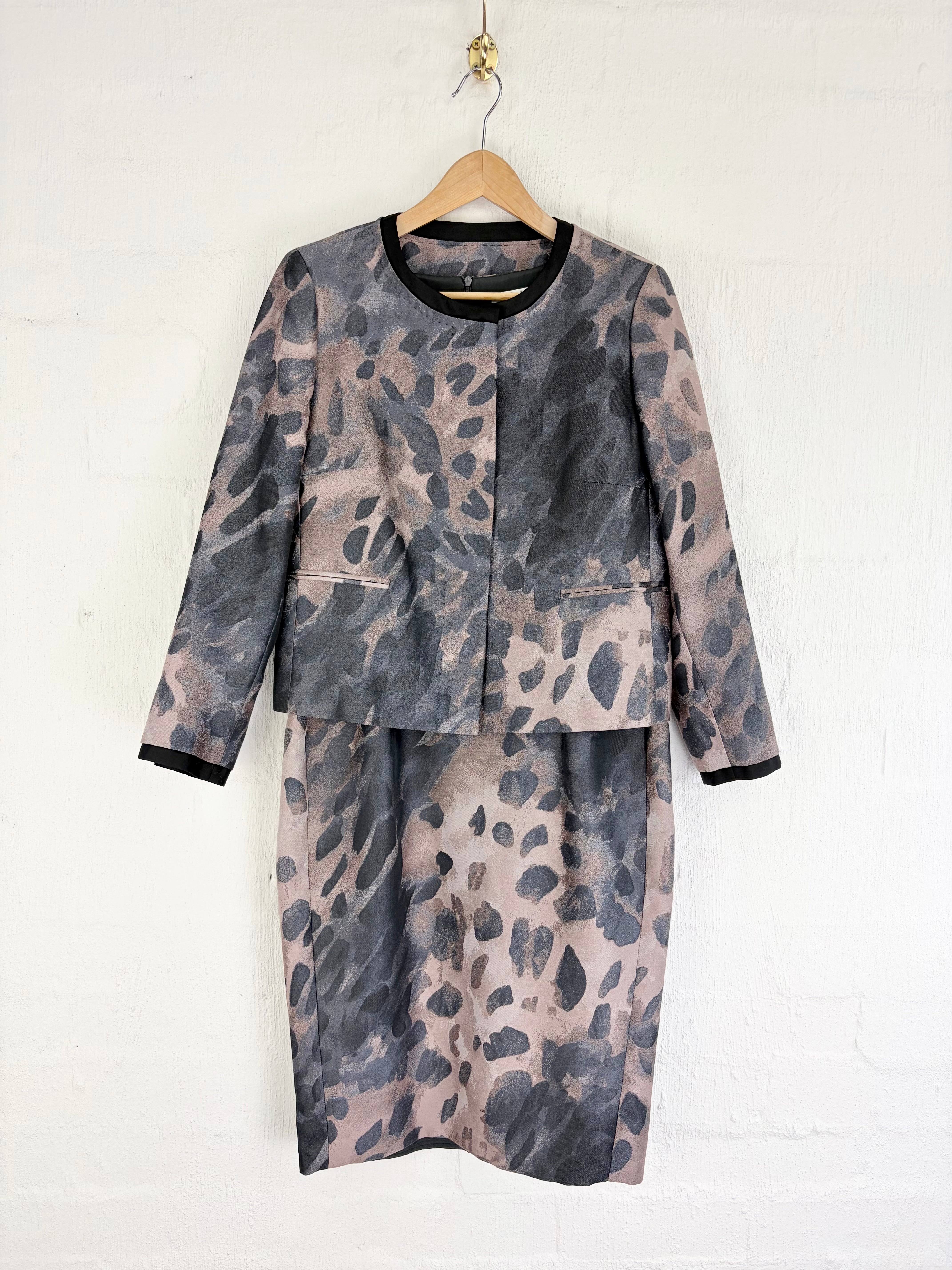 A two-piece skirt suit featuring a cropped jacket and matching pencil skirt in a gray and beige leopard print, hanging on a wooden hanger against a white brick wall.