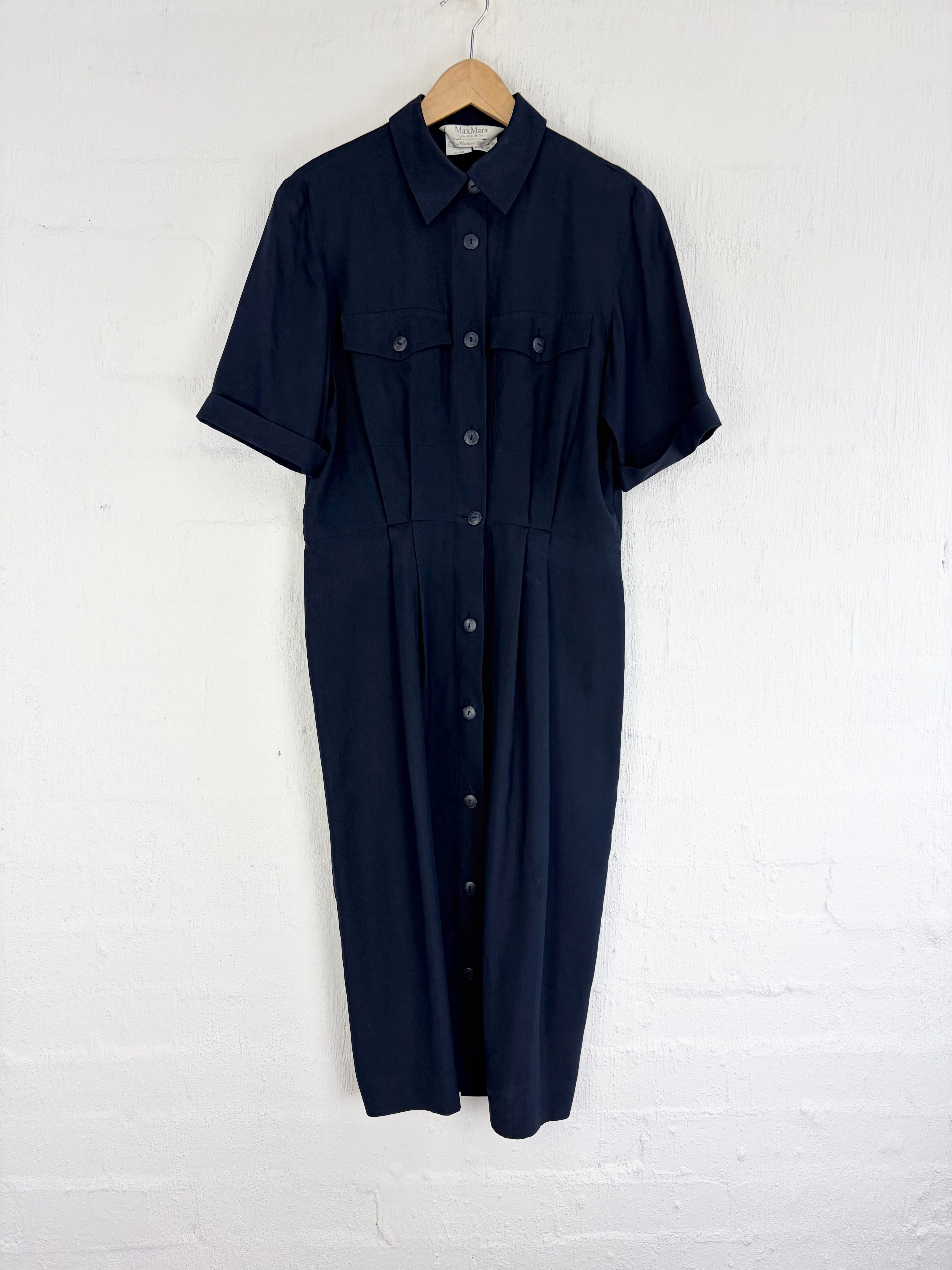 A dark navy blue short-sleeve button-front shirt dress with a pointed collar and chest flap pockets, hanging on a wooden hanger against a white painted brick wall.
