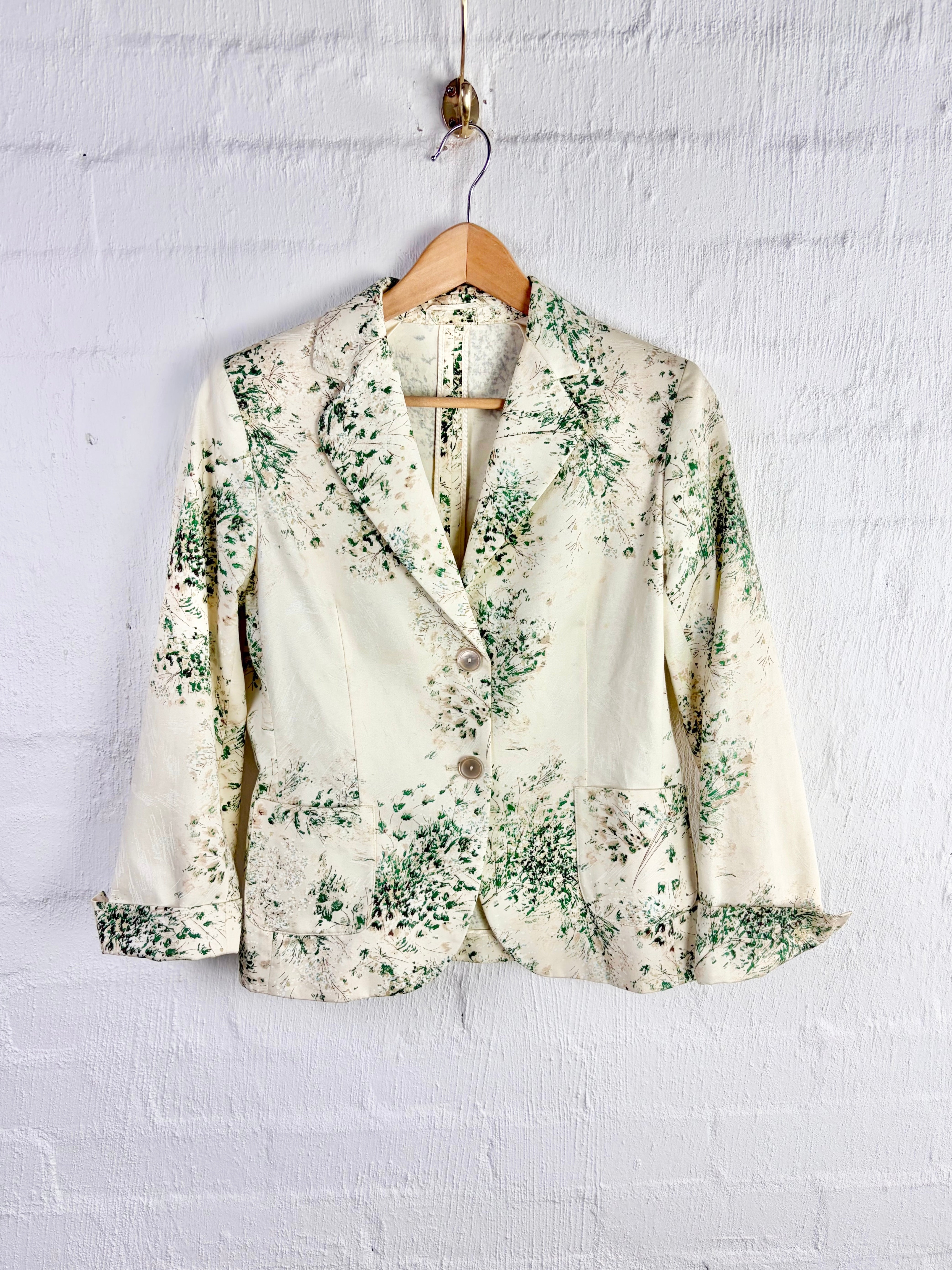 A cream-colored blazer with a green floral pattern, hanging on a wooden hanger against a white painted brick wall.
