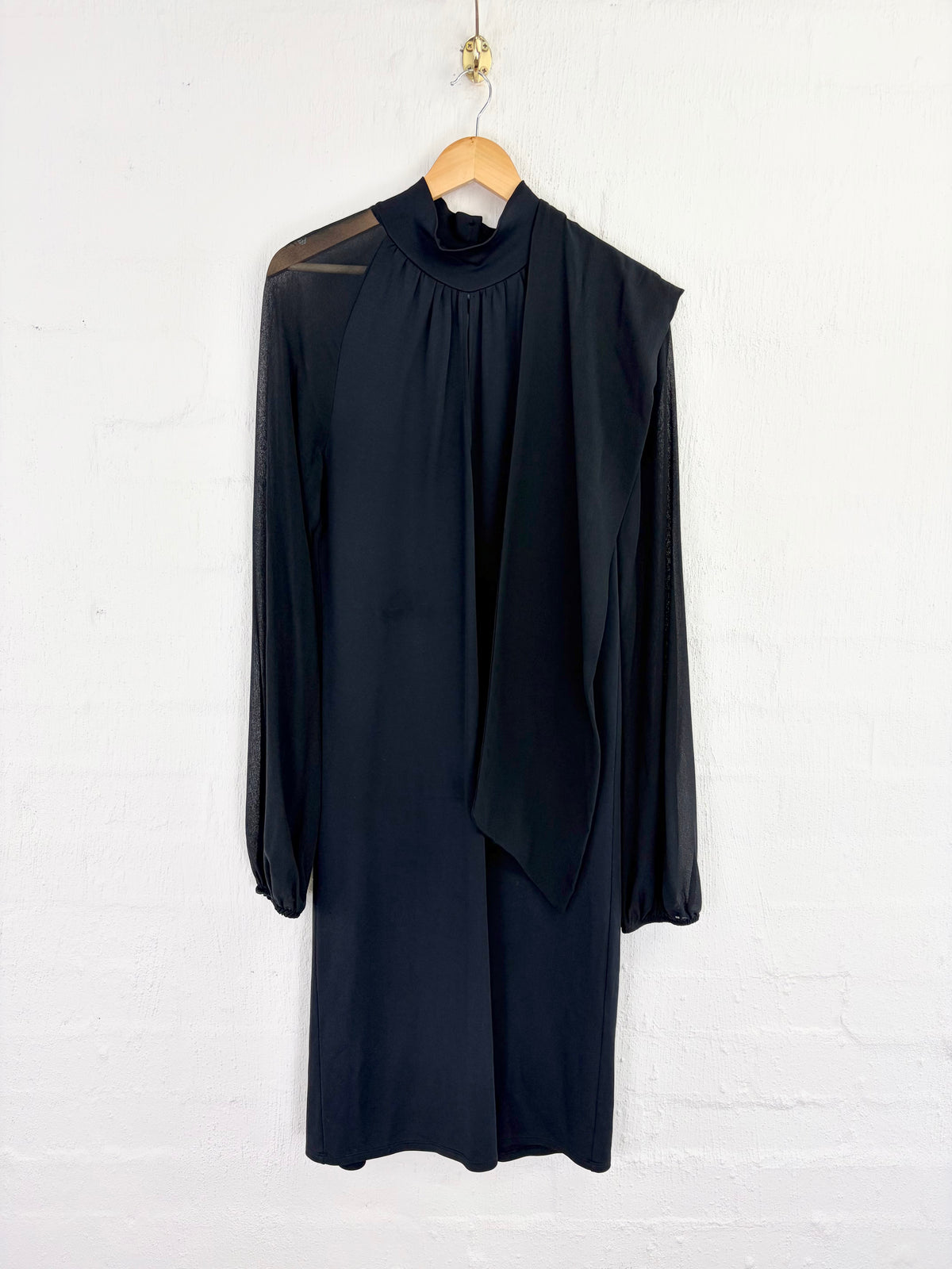 A black long-sleeve midi dress with a mock neck and draped tie detail, hanging on a wooden hanger against a white brick wall.