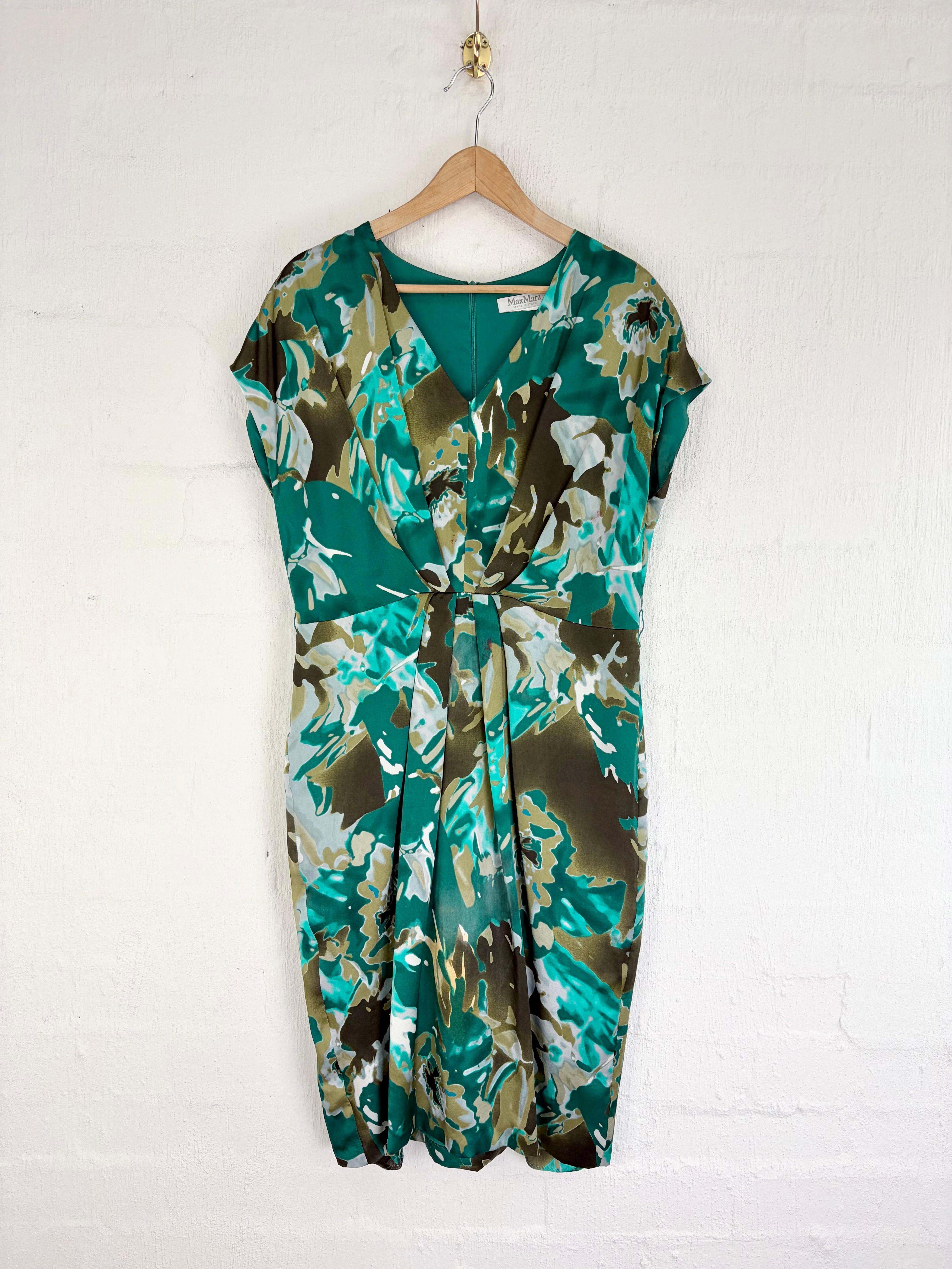 A short-sleeve wrap dress with a green, brown, and beige floral pattern, hanging on a wooden hanger against a white painted brick wall.