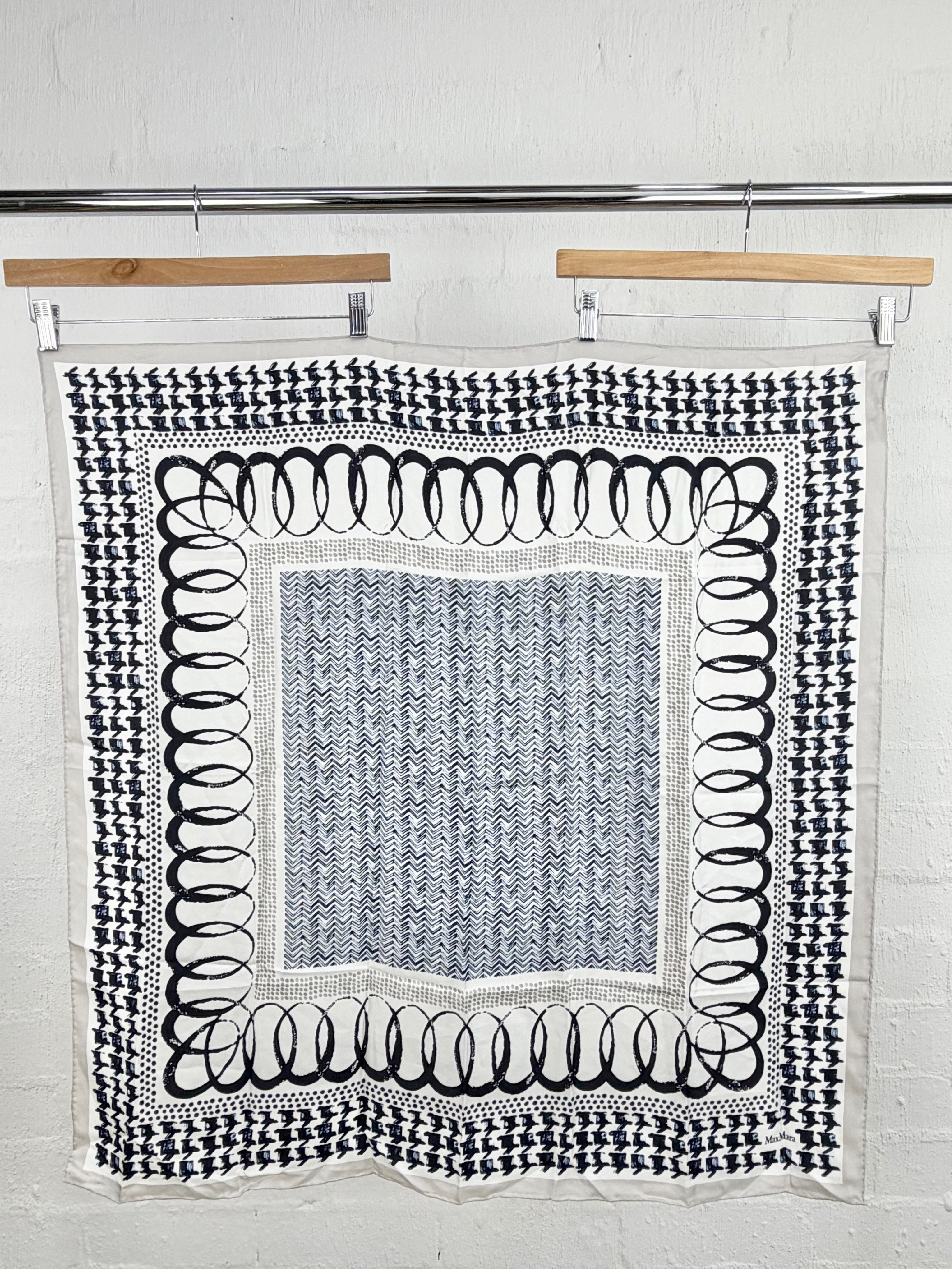 A square black and white patterned textile hanging from a wooden hanger on a metal rod against a white brick wall. The fabric features a central blue-gray patterned square surrounded by a white border with black circular motifs, and a black-and-white geometric patterned border.