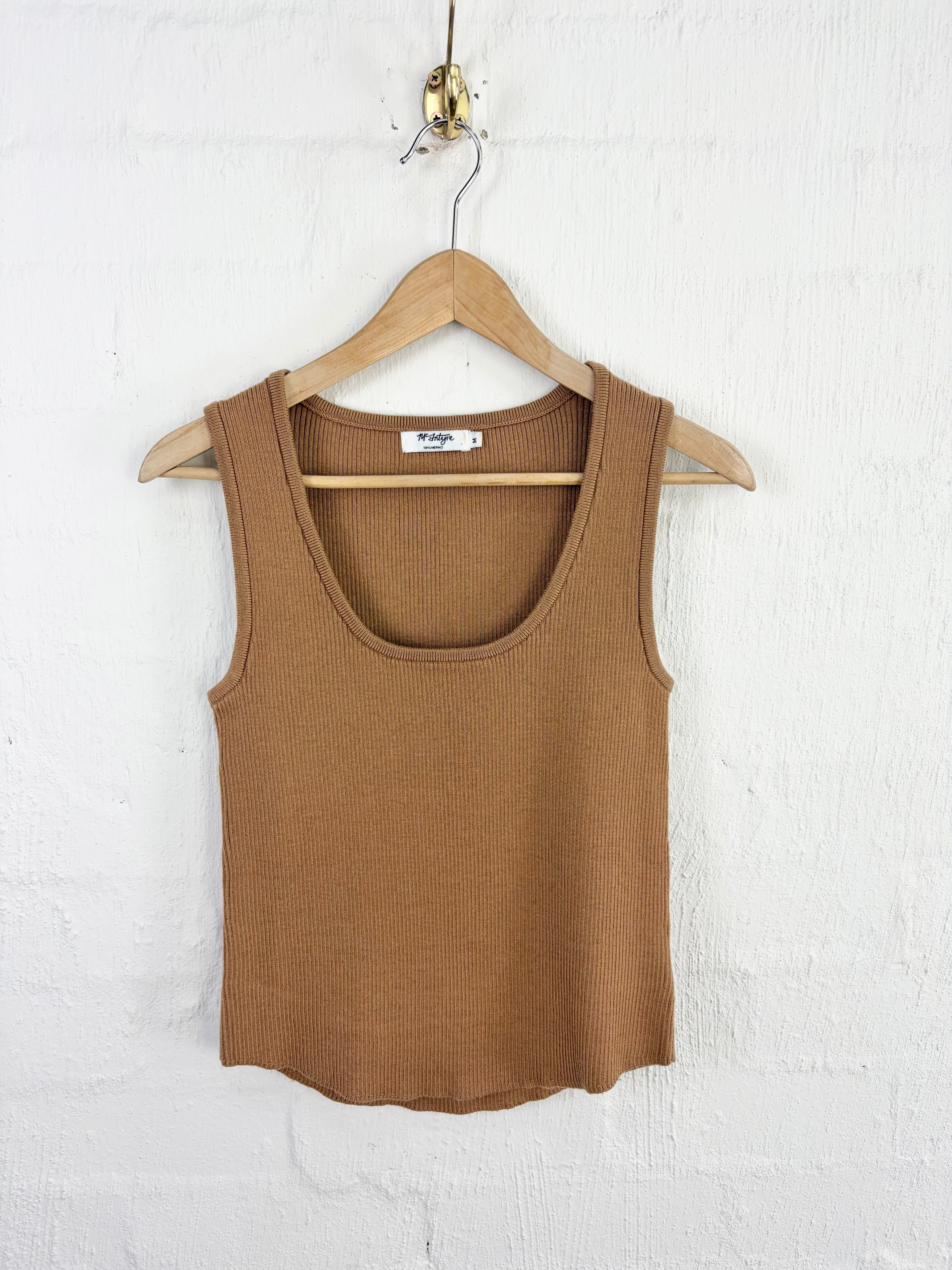 A ribbed knit scoop-neck tank top in a warm camel brown color, hanging on a wooden hanger against a white painted brick wall.