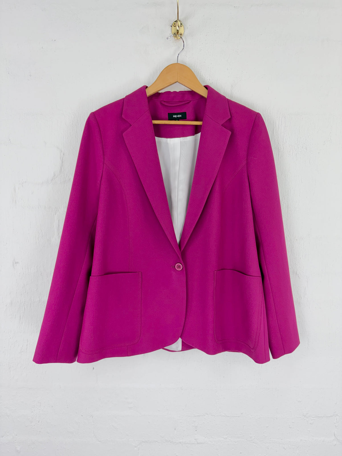 A bright magenta pink blazer jacket hanging on a wooden hanger against a white painted brick wall. The blazer features a single button closure, notched lapels, and two front patch pockets.