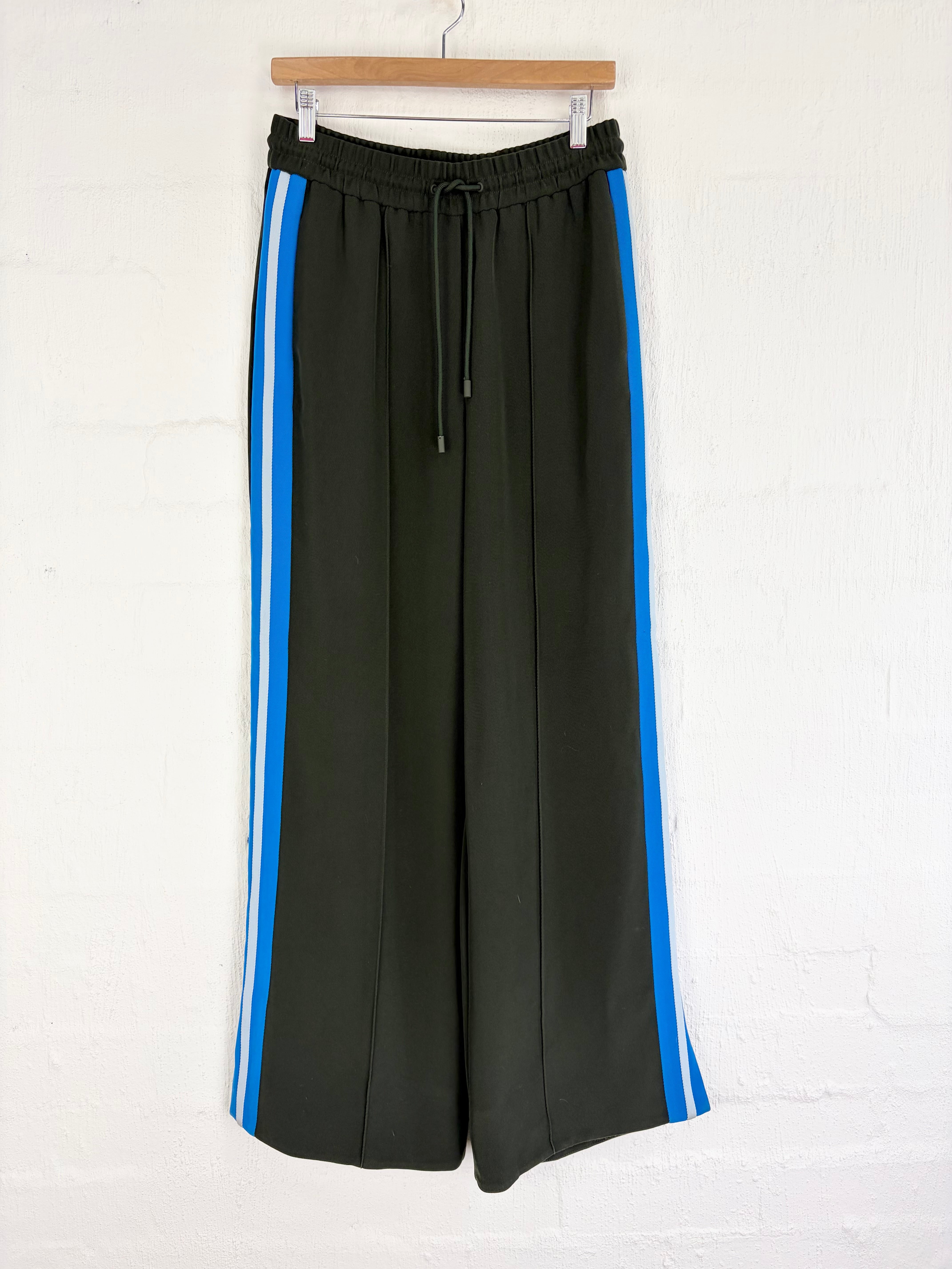 Black wide-leg cropped pants with bright blue side stripes, hanging on a wooden hanger against a white brick wall.